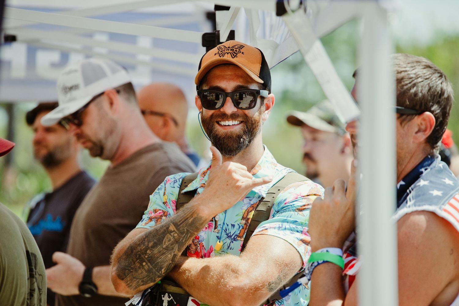 Smiling bearded man in floral shirt and cap giving shaka sign at outdoor event.