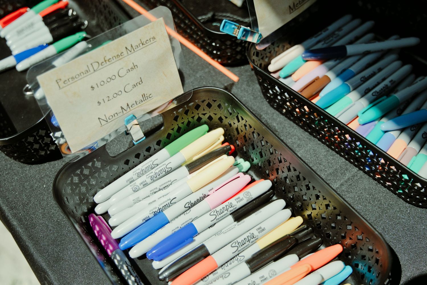 Colorful Sharpie markers organized in black mesh baskets labeled Personal Defense Markers at a vendor booth.