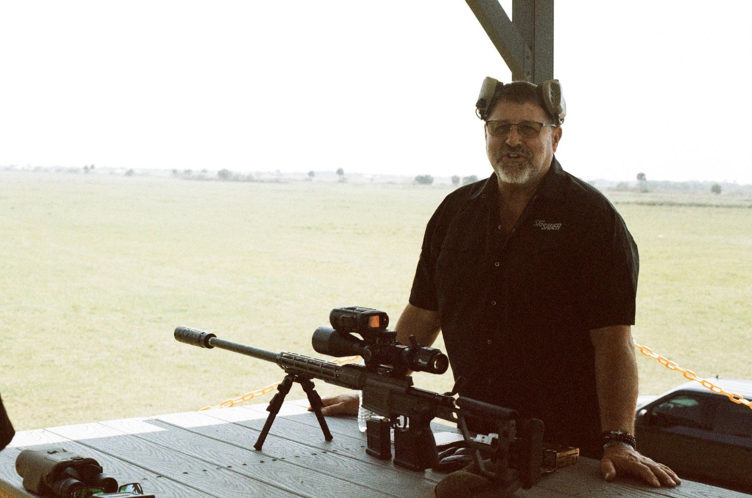 Man in black shirt stands behind a precision rifle with scope and suppressor on a table at an outdoor range.