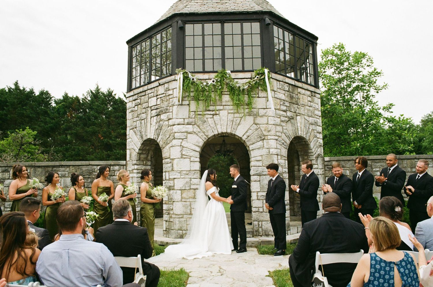 Outdoor wedding ceremony in front of a stone tower with ivy, bride and groom at altar, guests seated on white chairs.