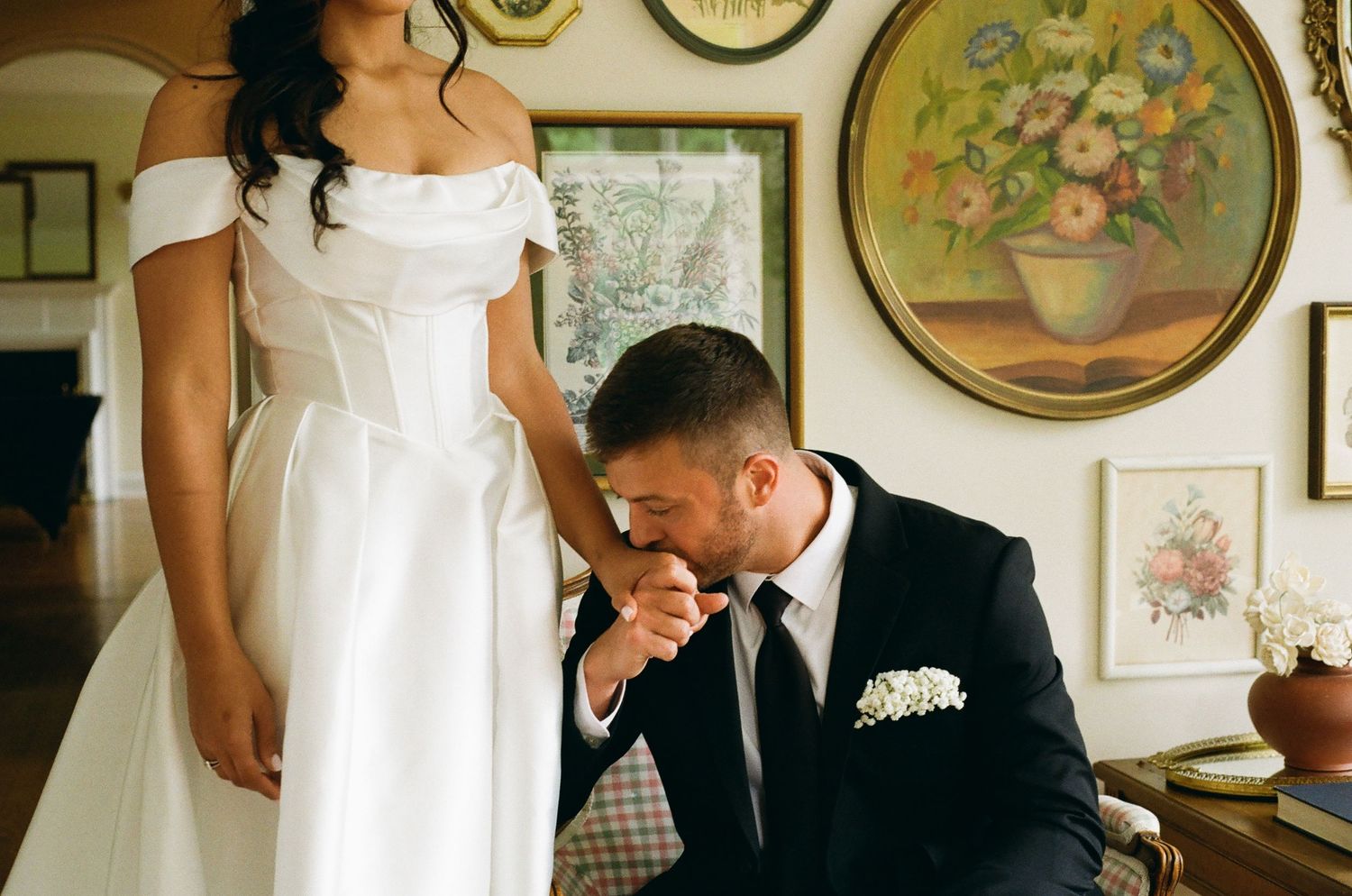 Groom in black suit kisses bride's hand during emotional wedding moment indoors with floral paintings on wall.