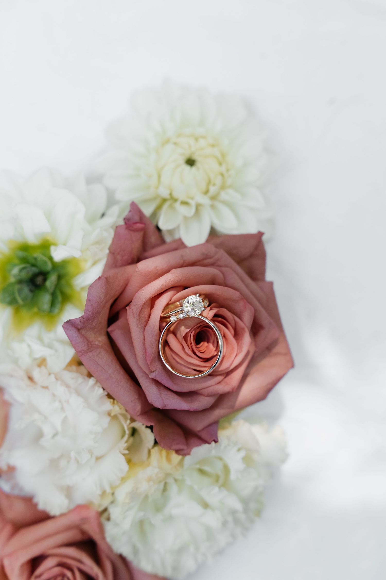 Wedding rings nestled in a dusty pink rose surrounded by white carnations and dahlias in a bridal bouquet.