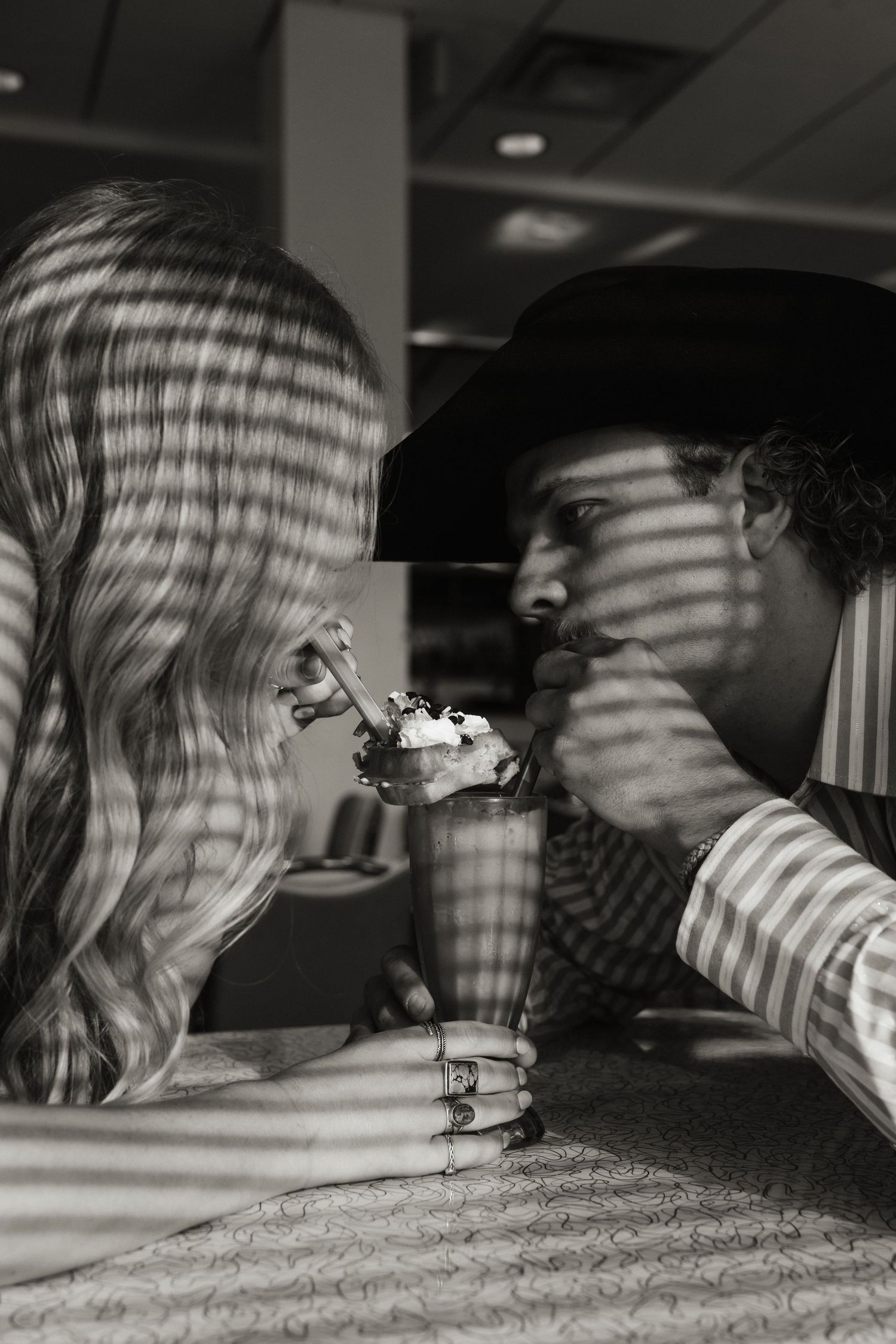 Black and white photo of a couple sharing a milkshake at a diner, leaning close together romantically.