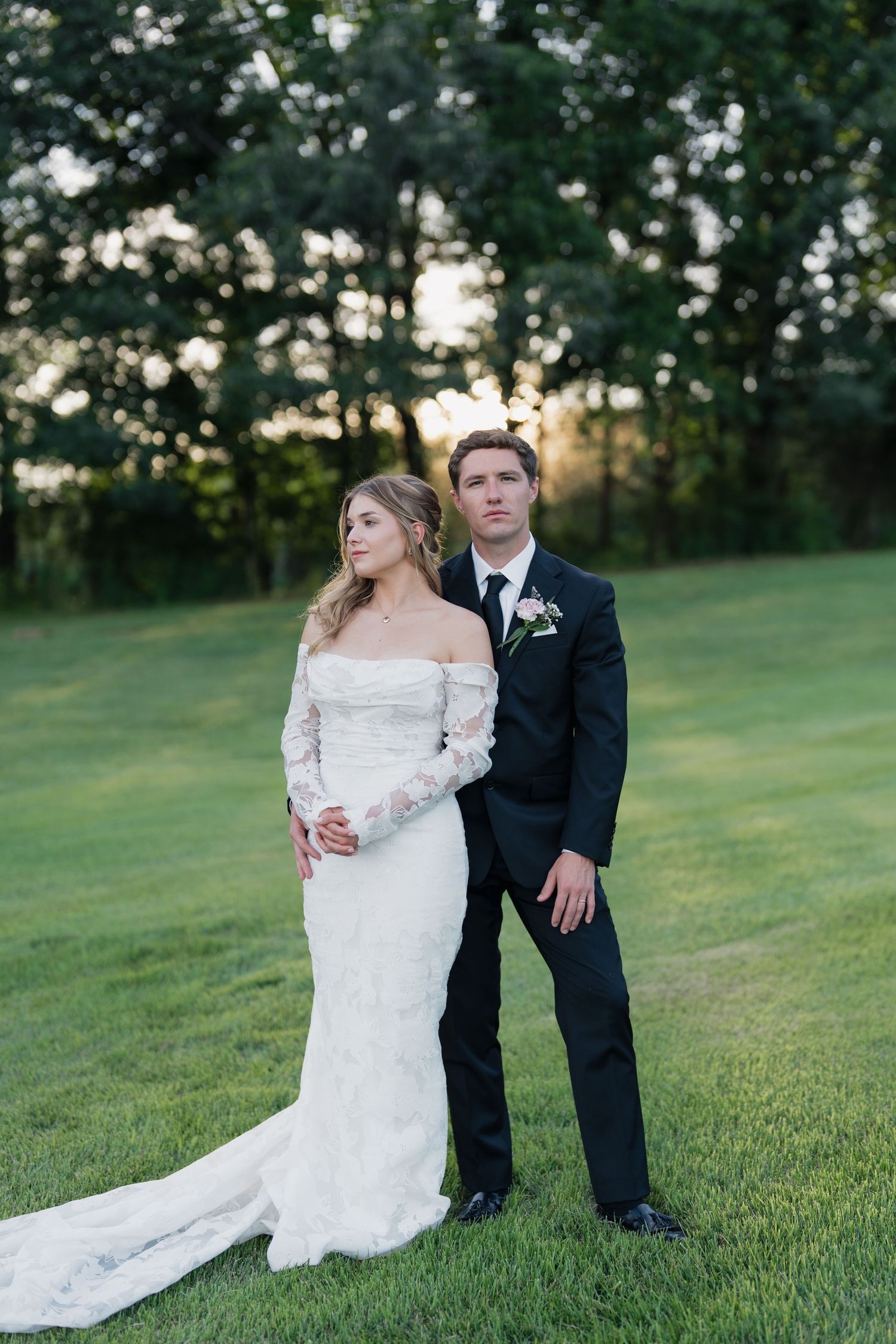 Bride in lace off-shoulder gown and groom in black tuxedo pose on green lawn at golden hour.