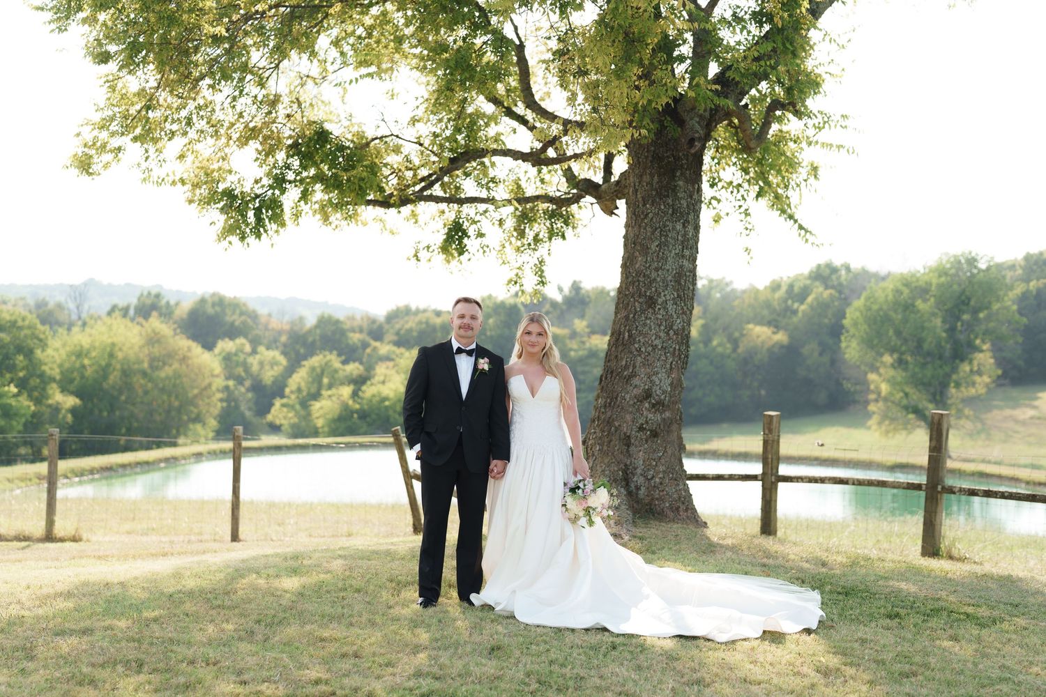 Bride and groom posing under a large tree with a scenic pond and rolling hills in the background.