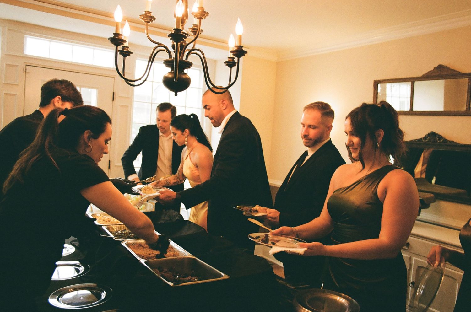 Elegantly dressed guests serving themselves at a buffet table under a chandelier at a formal wedding reception.