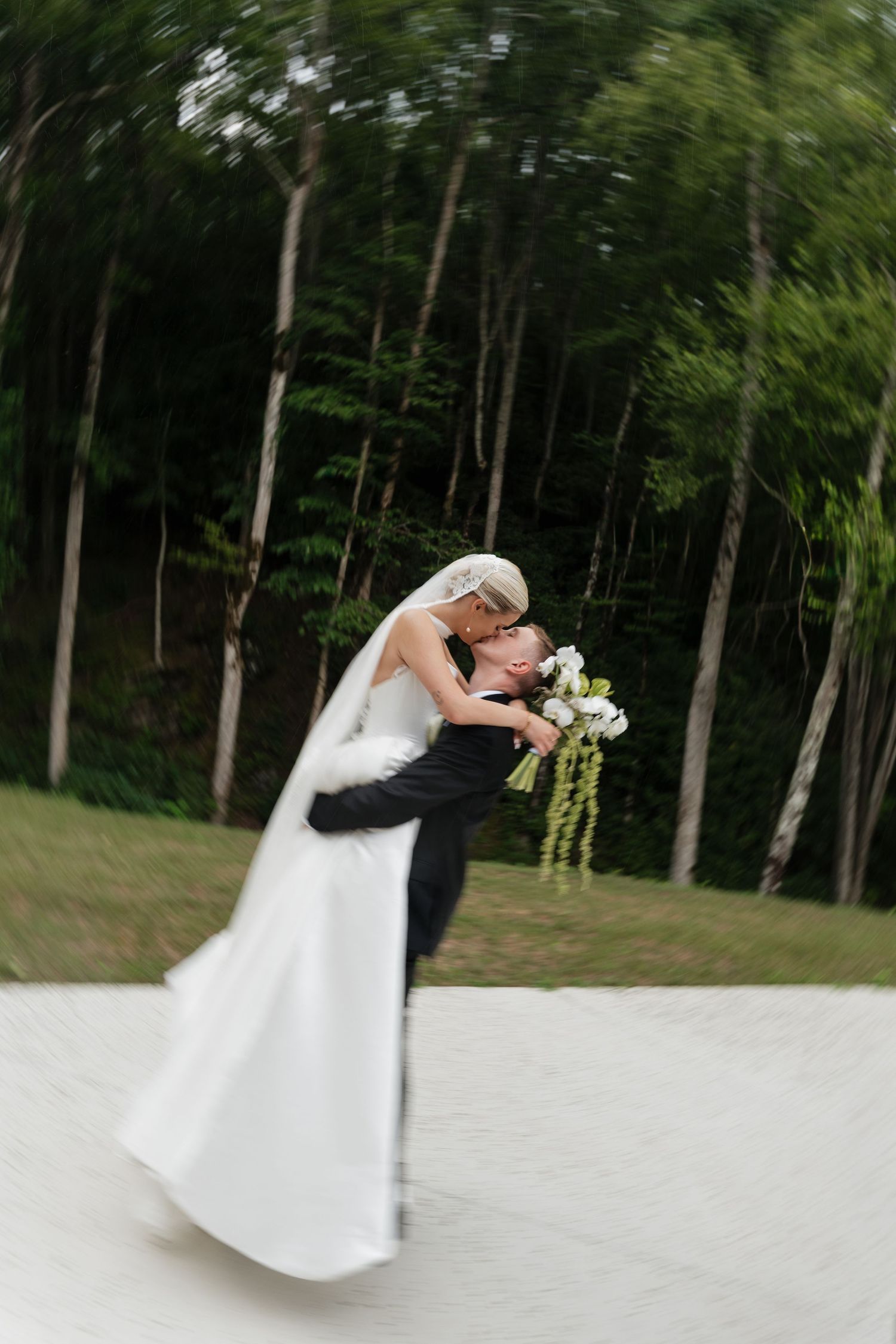 Bride and groom share a romantic dip kiss outdoors, surrounded by lush green trees, with motion blur capturing the joyful moment.