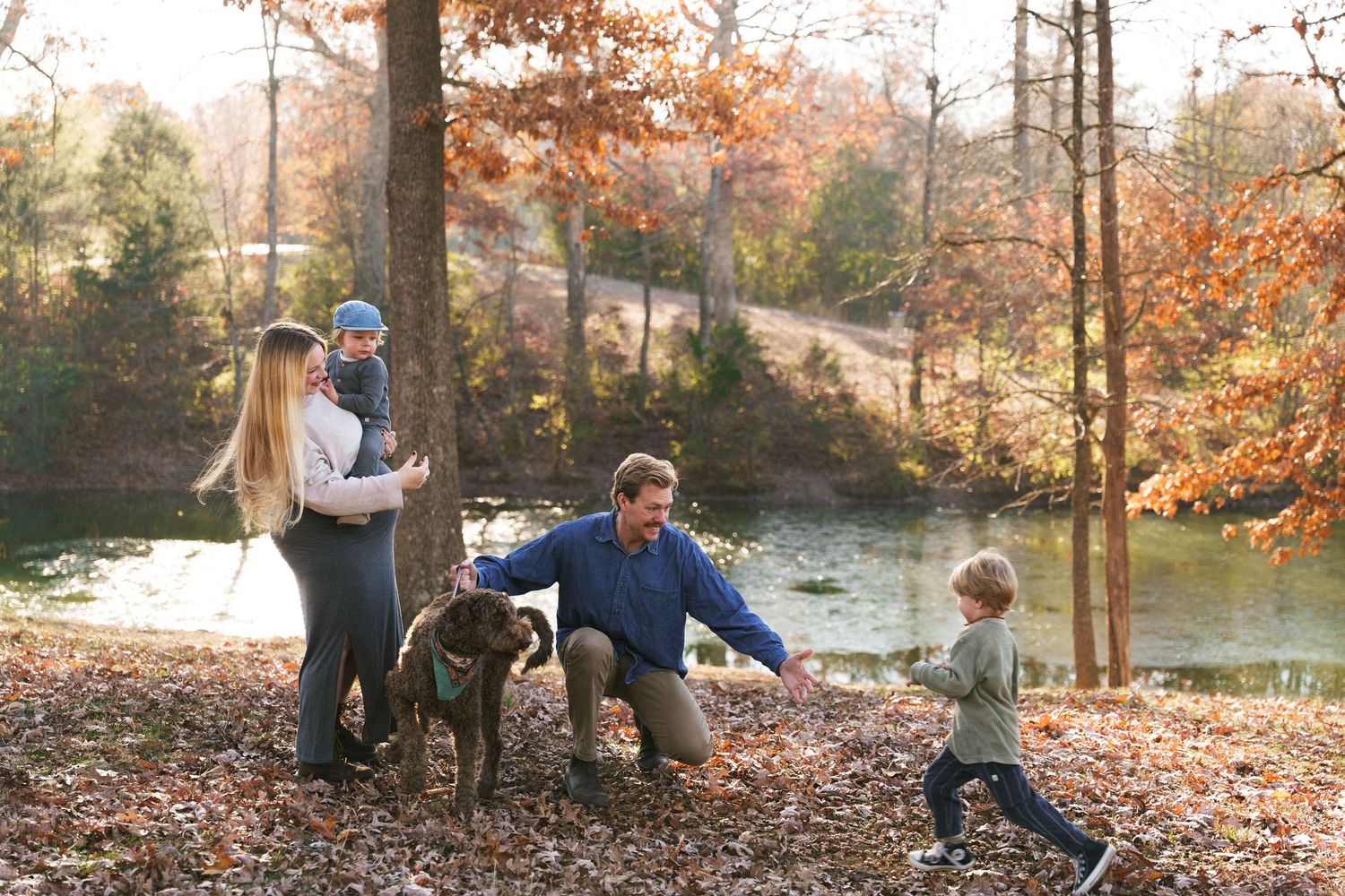 Family enjoys autumn day at lakeside park with their dog as colorful fall foliage surrounds them in golden light.