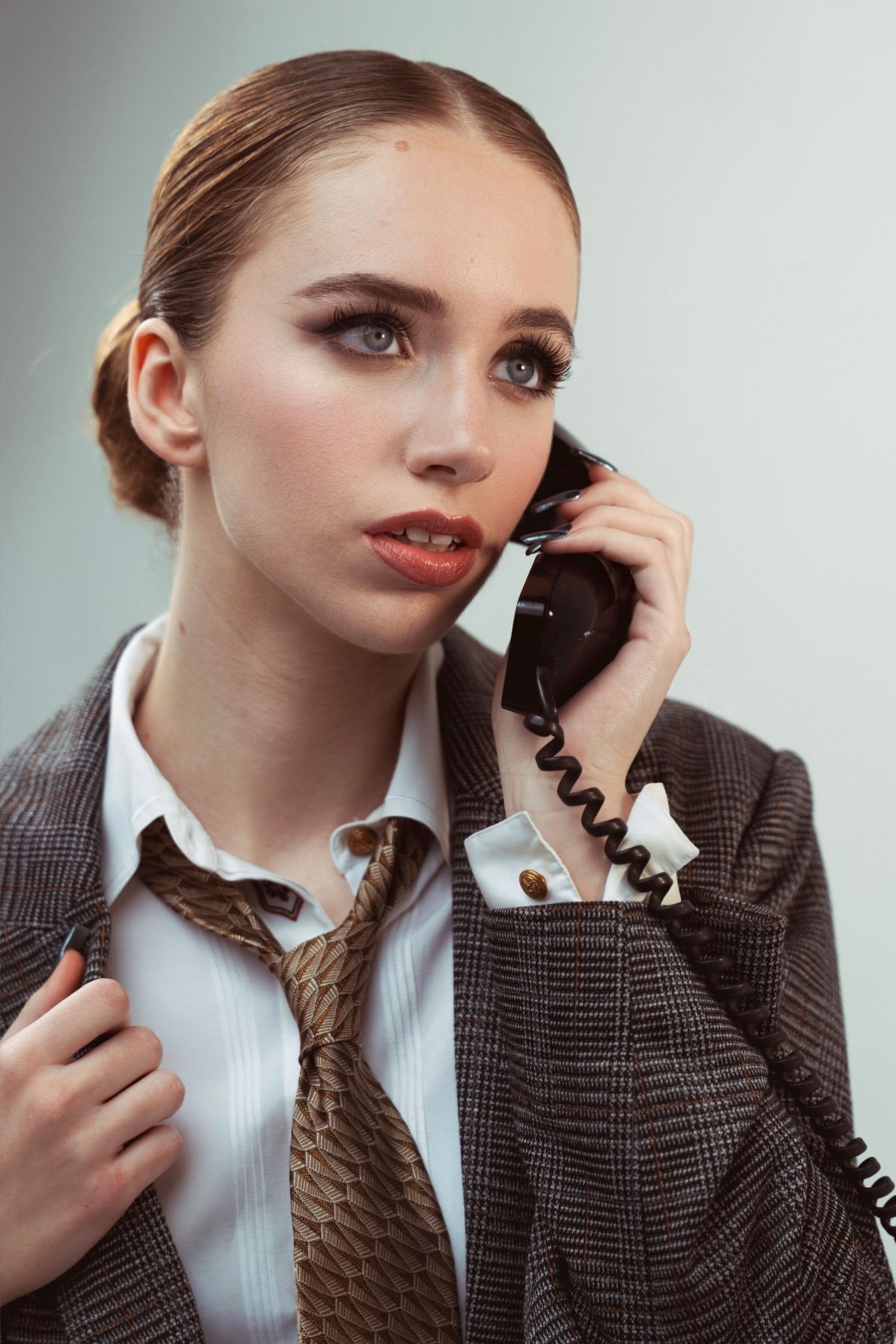 A professional wearing a brown tweed blazer and white blouse speaks on a vintage telephone with red lipstick and slicked back hair.