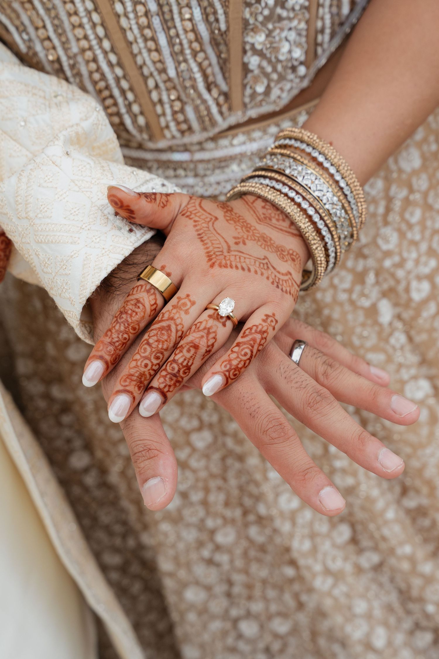 Close up of intricate henna design on hand with white nail polish and sparkly bangles against embellished fabric.