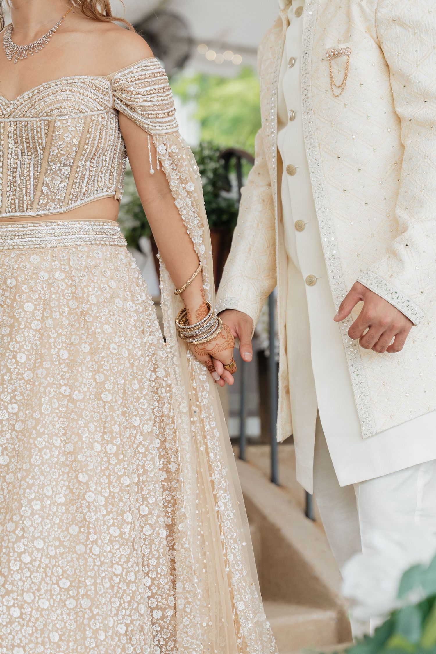 Wedding couple holds hands showcasing intricate beaded details on their matching white and cream colored outfits.
