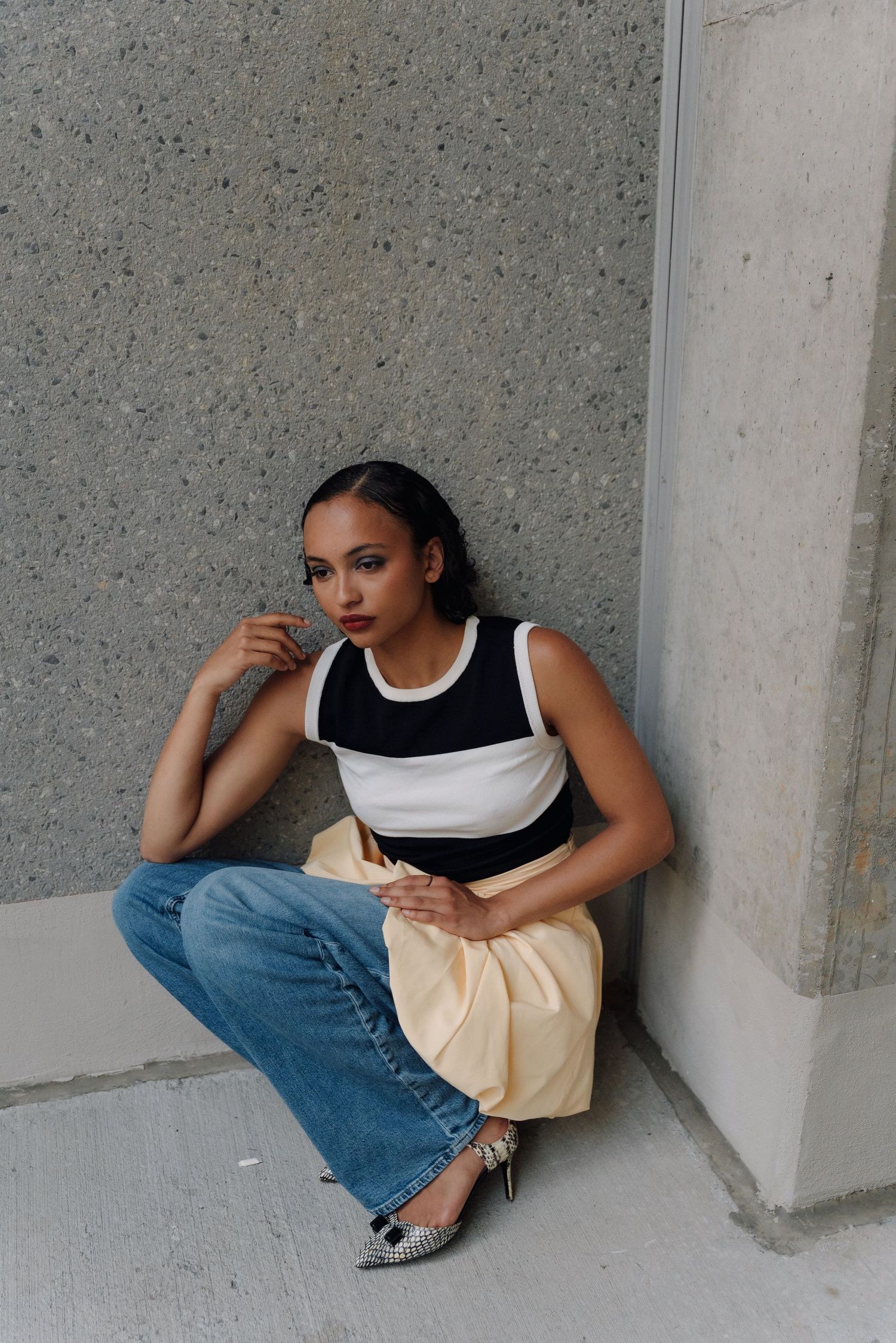 Young woman in striped crop top, yellow skirt, and jeans sits against a concrete wall outdoors.