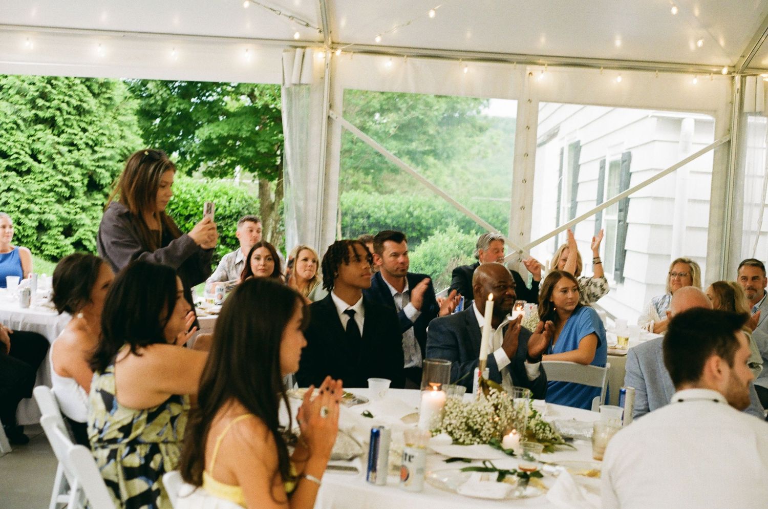 Wedding reception toast under a tent with guests applauding as a woman speaks at an elegant outdoor celebration.