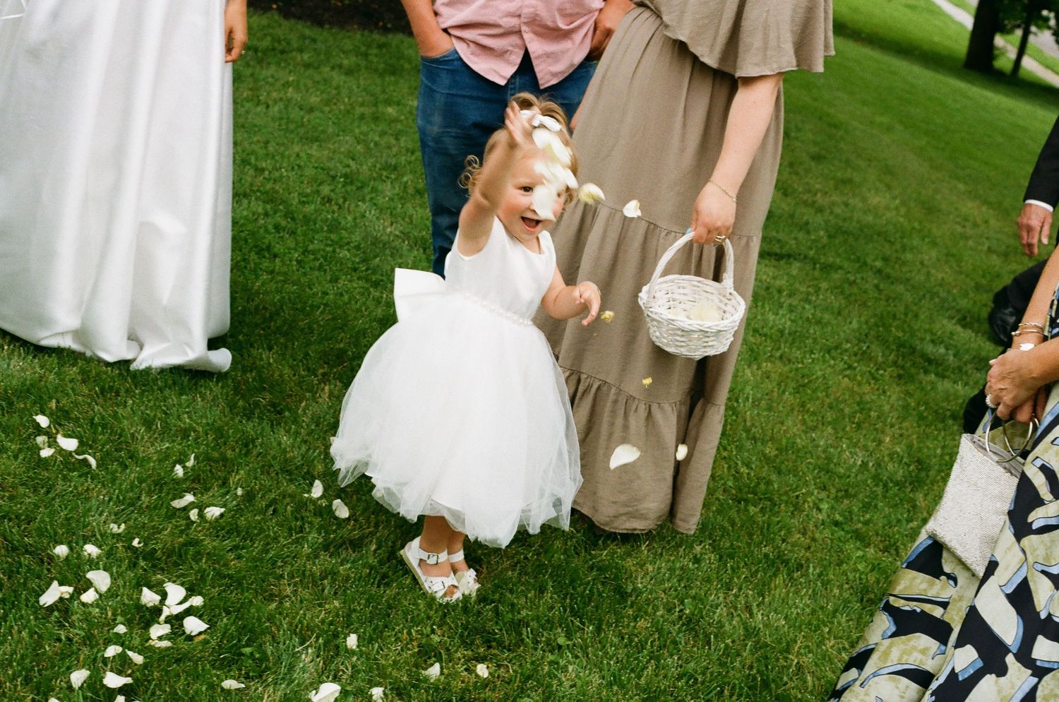Flower girl in white tutu dress scattering rose petals on green grass during outdoor wedding ceremony.