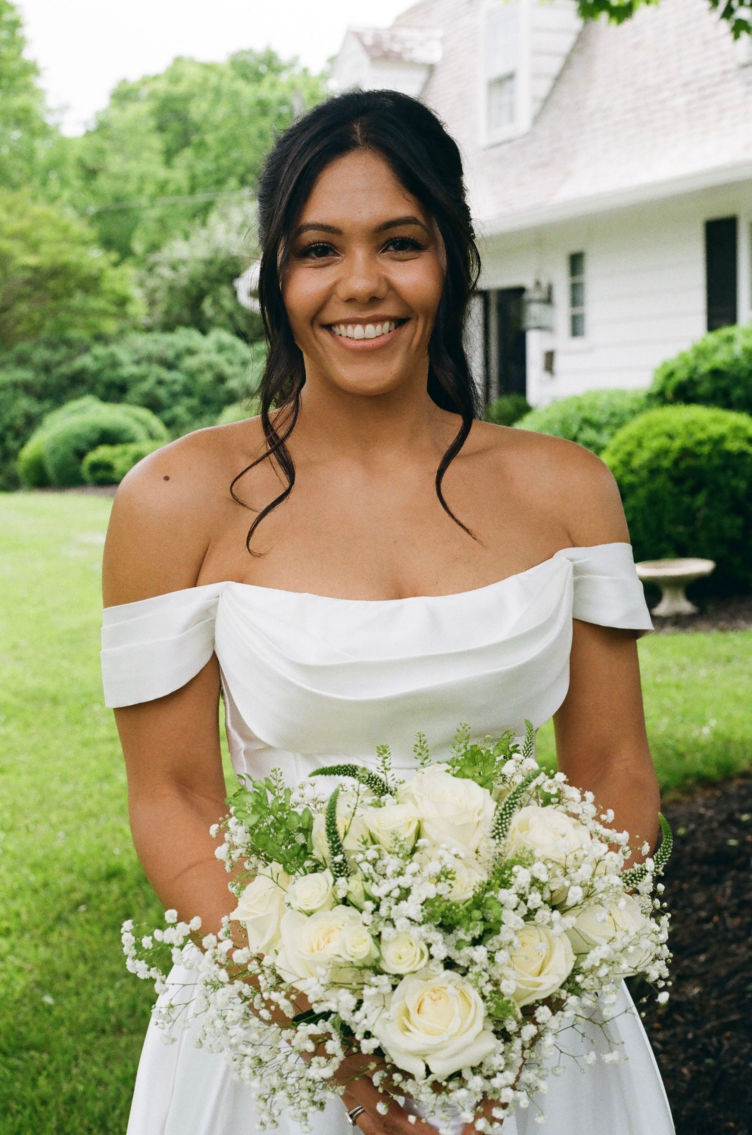 Smiling bride in off-shoulder white gown holds bouquet of white roses and baby's breath outdoors.