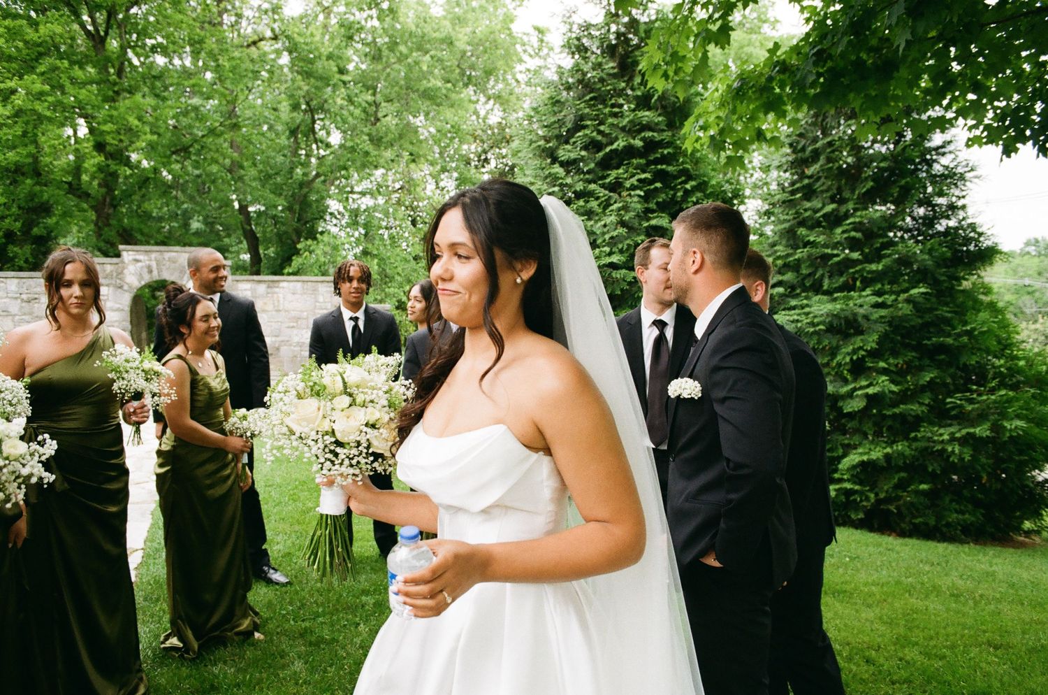 Bride in strapless white gown and veil smiles outdoors with bridesmaids in green dresses and groomsmen behind her.