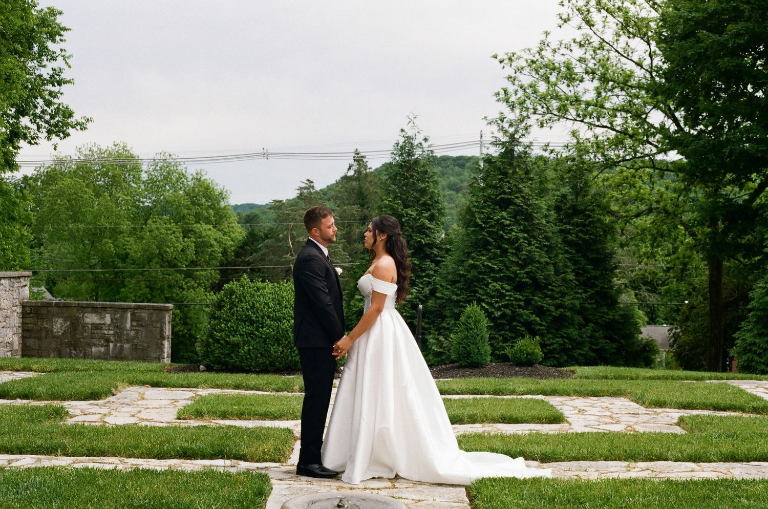 Bride and groom hold hands in lush garden with bridge views on their wedding day.