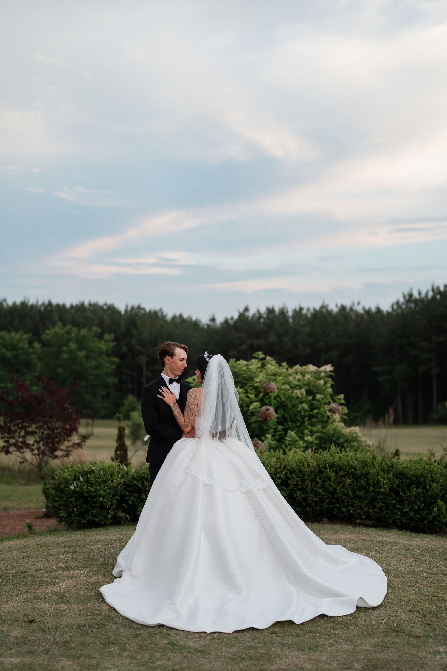 Bride in white gown with long veil and groom in black tuxedo stand together outdoors with lush green trees backdrop.