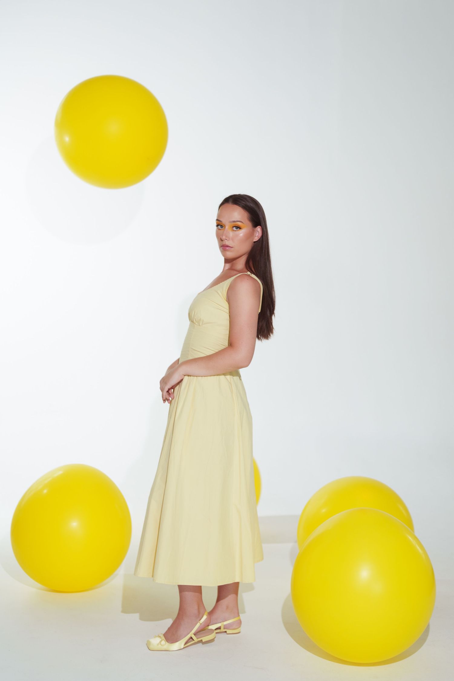 Woman in yellow midi dress poses with large yellow balloons in a bright white studio setting.