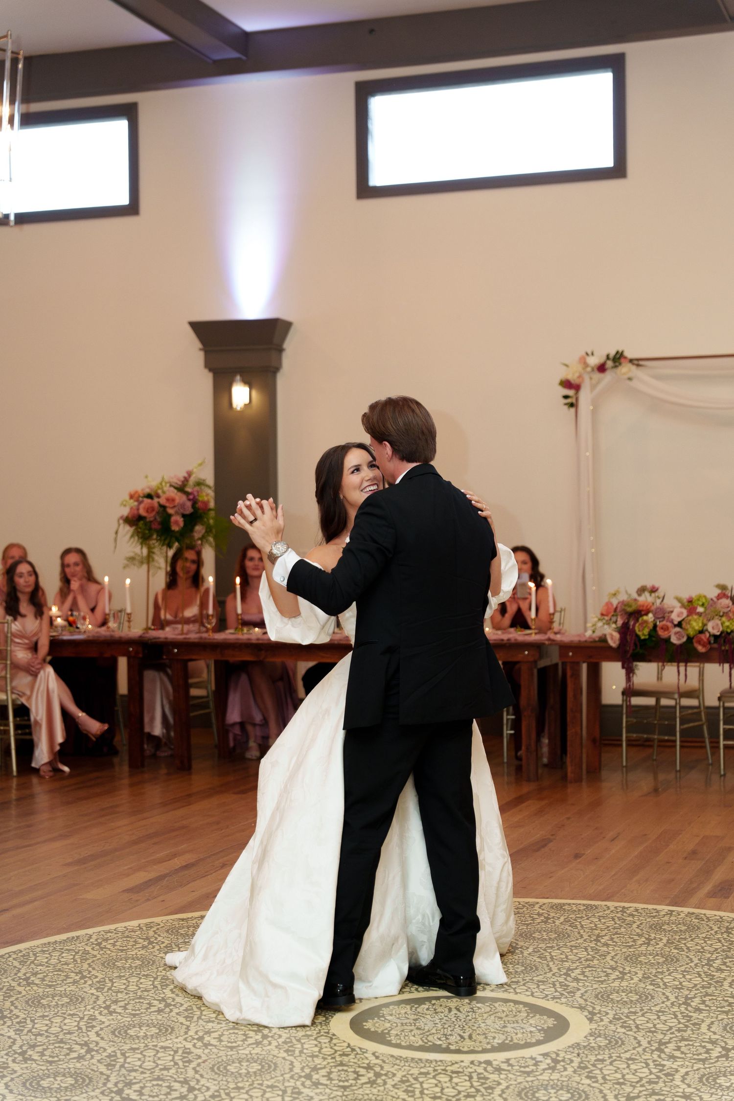 A couple shares their first dance at their wedding reception while guests watch from rustic wooden farm tables.