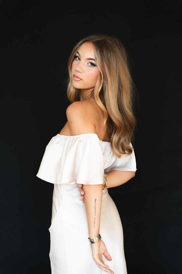 Young woman in white off-shoulder dress with tattoo and bracelet posing against black background.