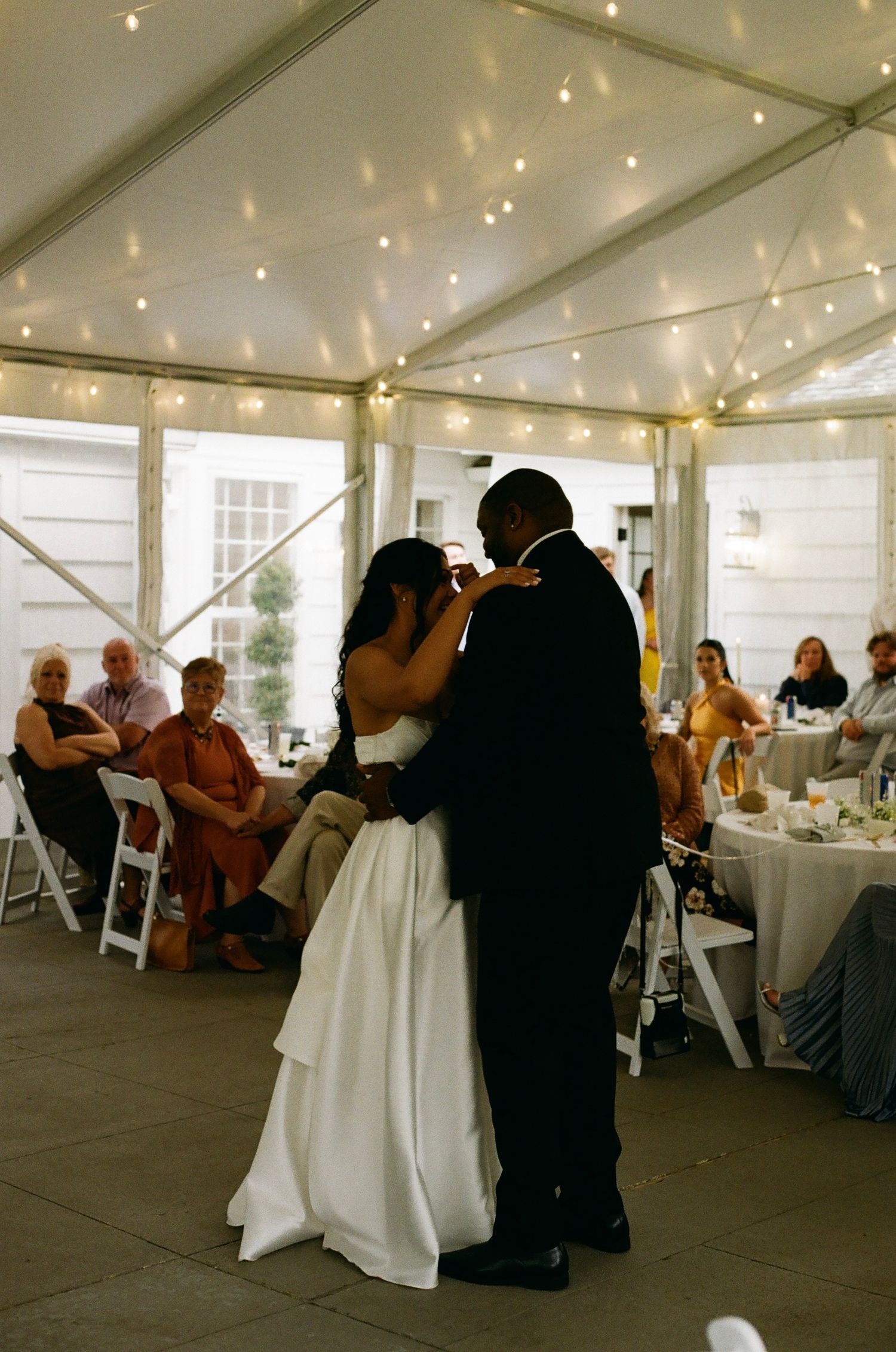 A bride and groom share their first dance under twinkling lights in a elegant wedding tent while guests look on.