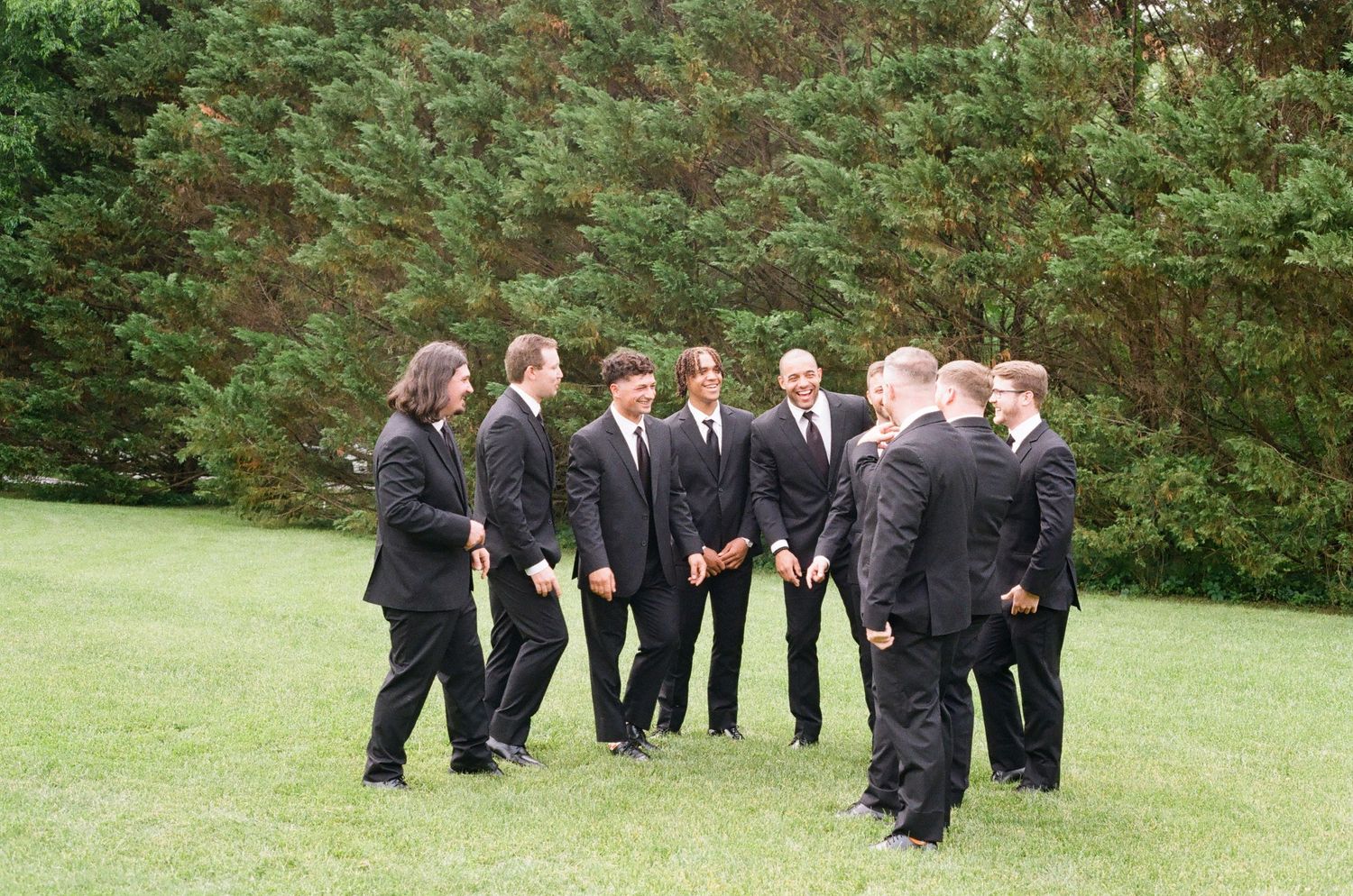 Groomsmen in black suits gathered on green grass with pine trees in background at outdoor wedding.