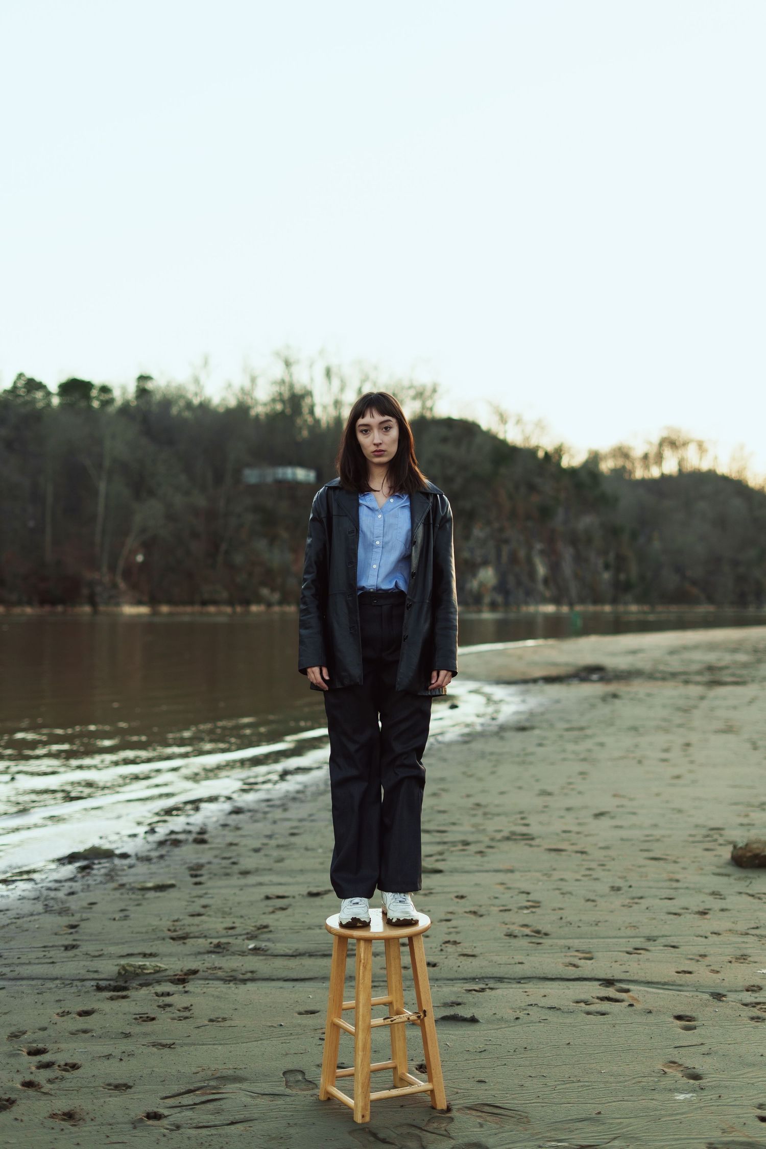 Person stands on wooden stool at beach wearing leather jacket and denim during sunset by calm water.