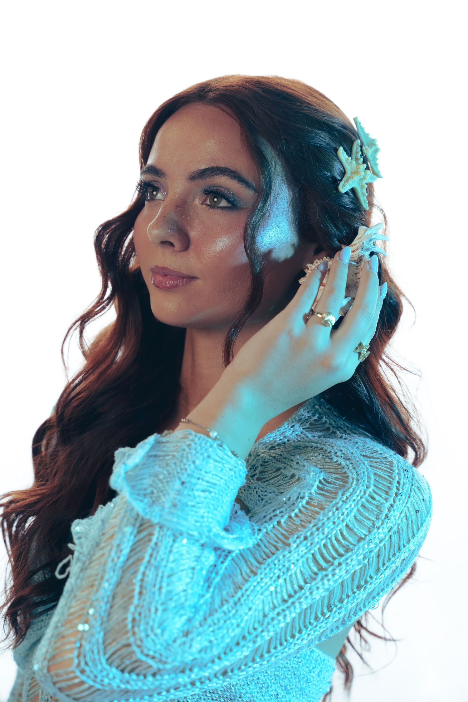 Portrait of a woman in blue lighting wearing a knit sweater and floral hair accessory, with long wavy dark hair and rings.