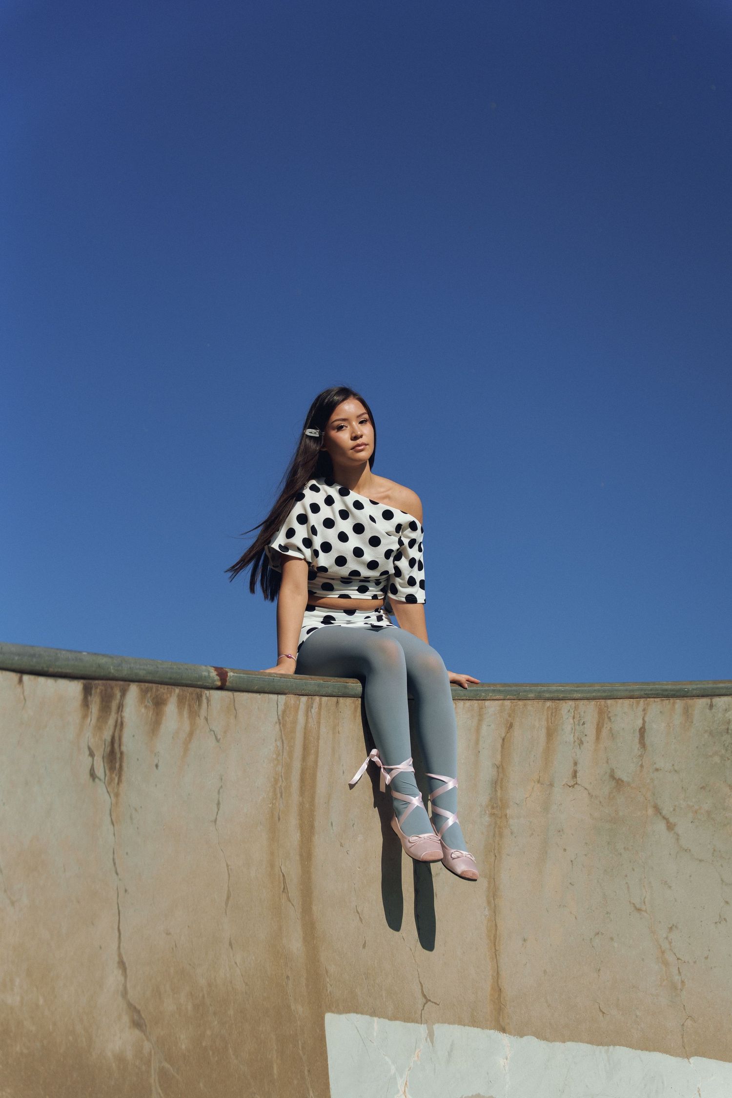 A person in a polka dot top and gray leggings sits on a concrete wall against a bright blue sky.
