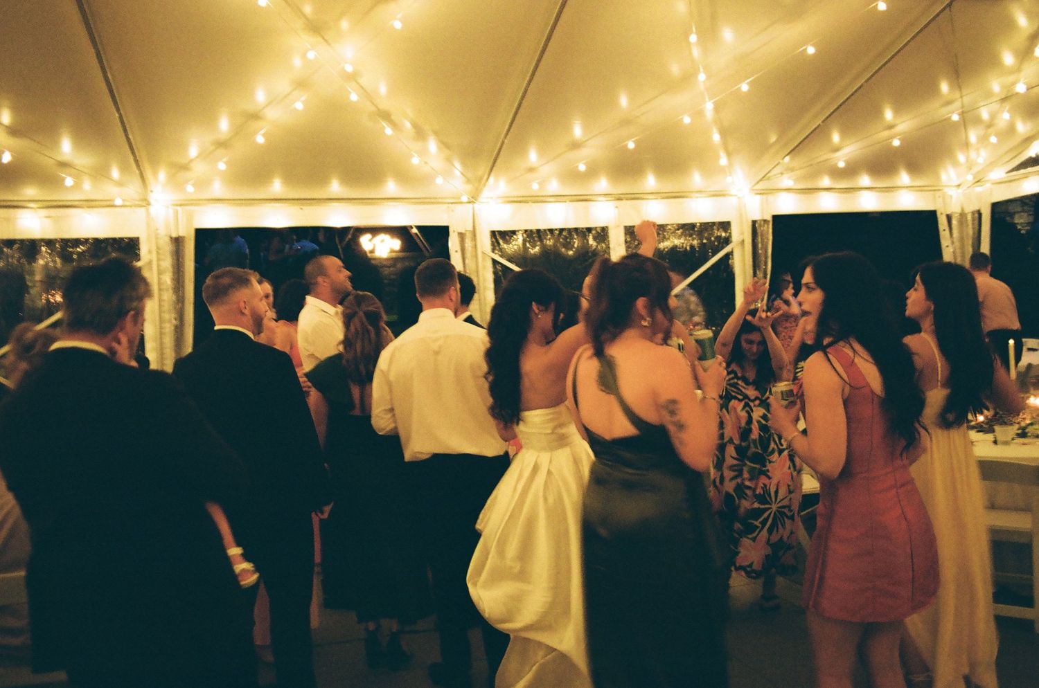 Guests dancing under warm string lights at an elegant outdoor wedding reception tent at night.