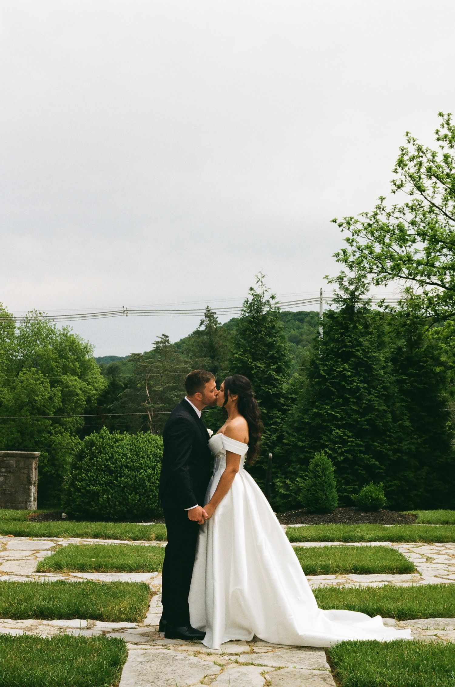Bride and groom share a kiss in a lush garden, she wears an off-shoulder white ballgown, he wears a black tuxedo.