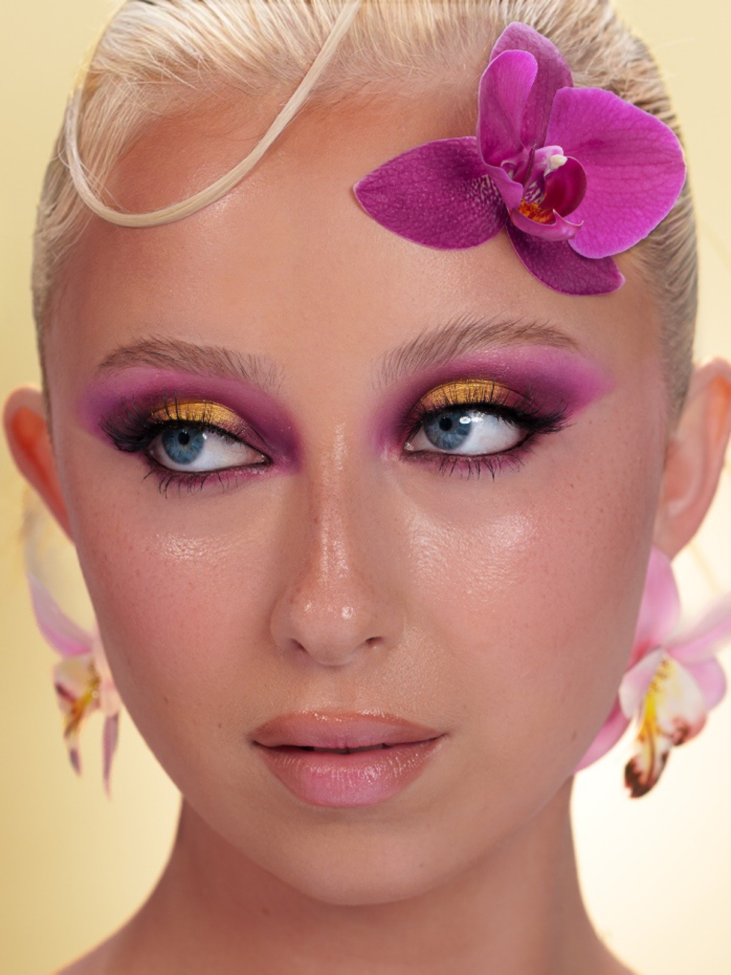 Close-up of a woman with bold pink and gold eye makeup, blue eyes, and a purple orchid flower in her blonde hair.