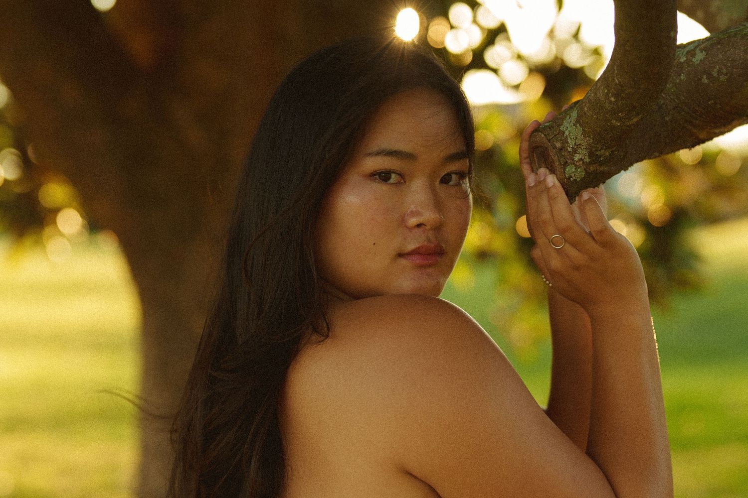 Young Asian woman posing gracefully by a tree in golden sunlight, looking over her shoulder outdoors.