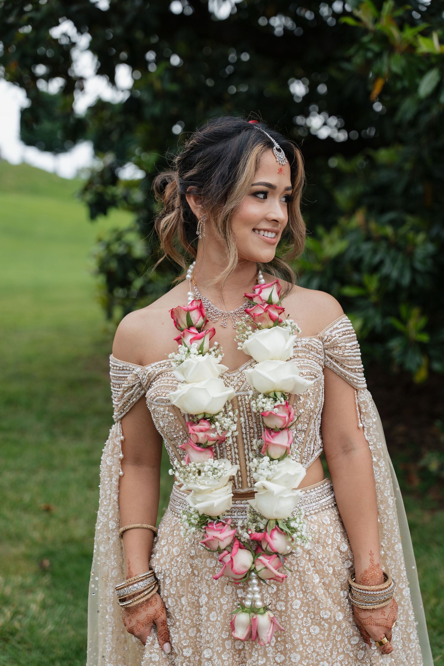 Woman in sparkly beige dress wearing floral garland at outdoor celebration.