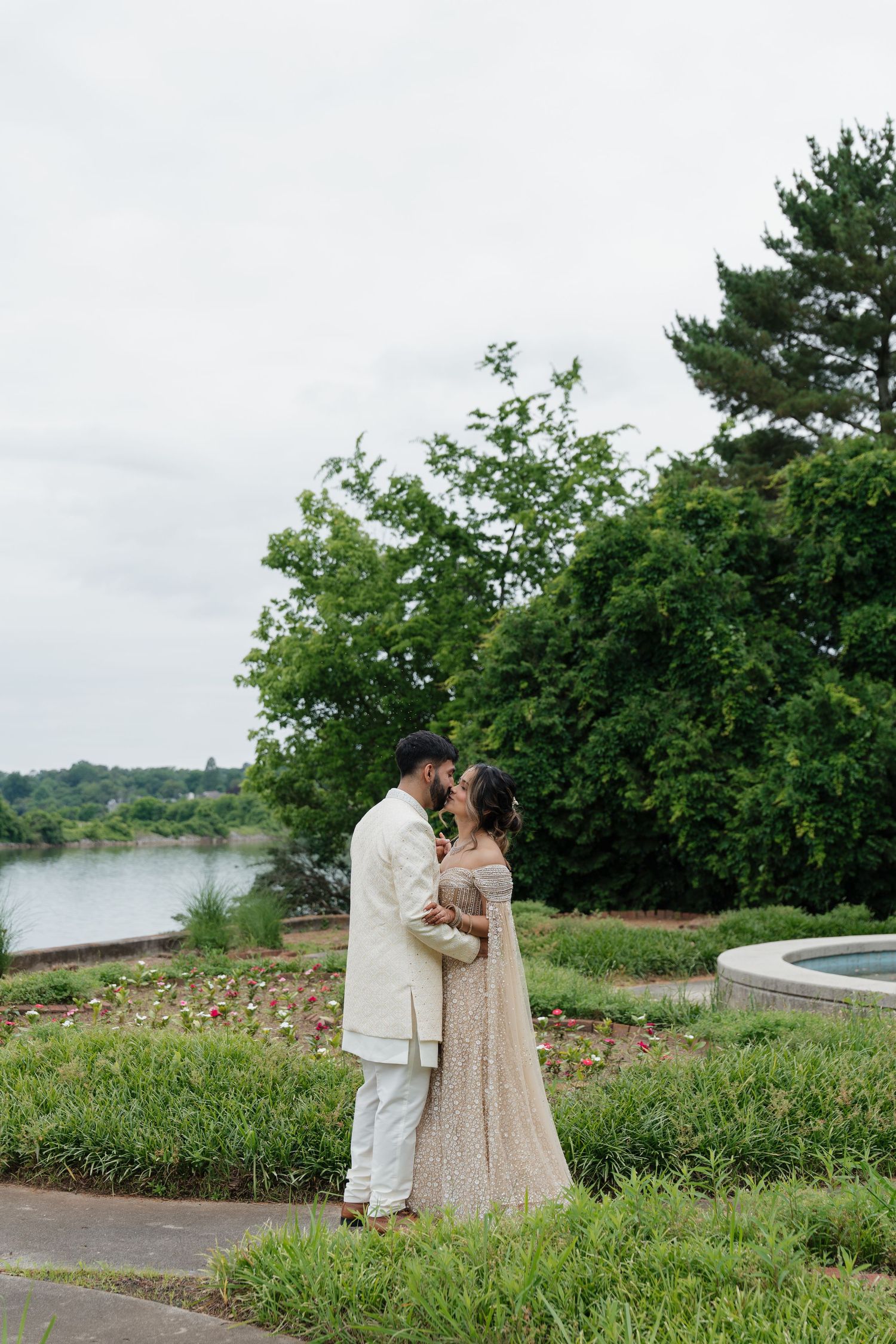 A couple shares a romantic moment on a grassy hill overlooking a scenic lake with green trees in the background.