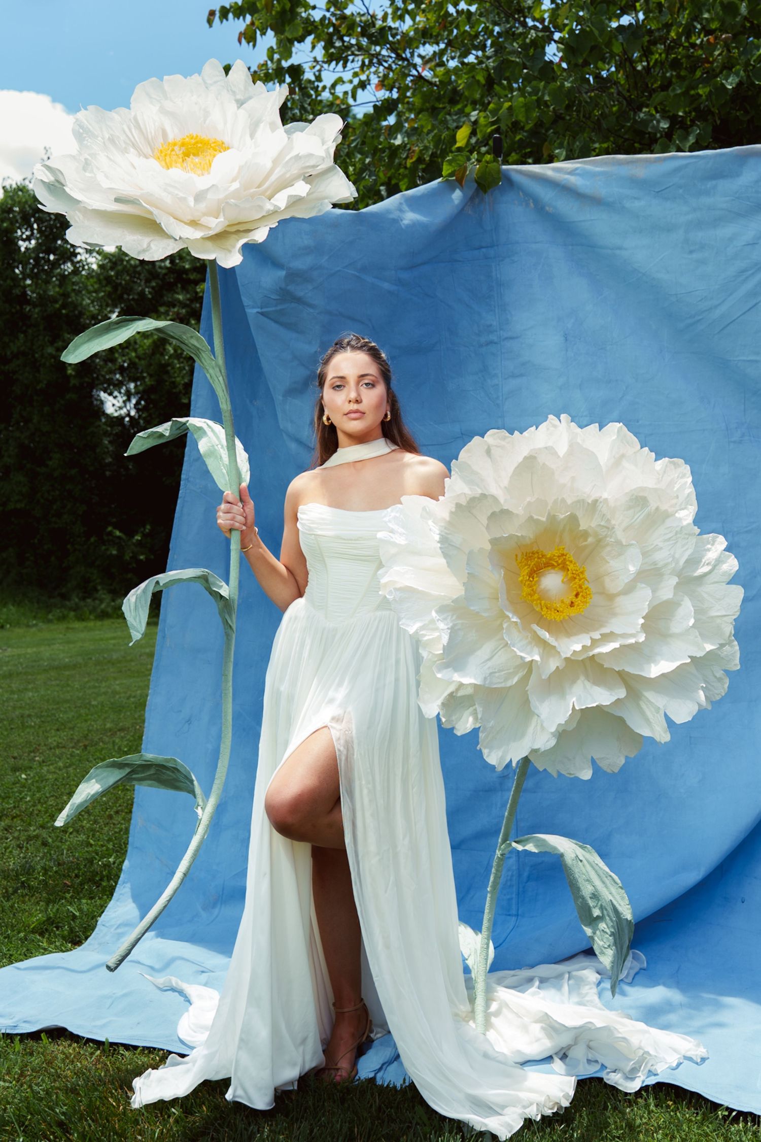 A fashion photography series with white flowy dresses and oversized paper flowers against a blue backdrop.