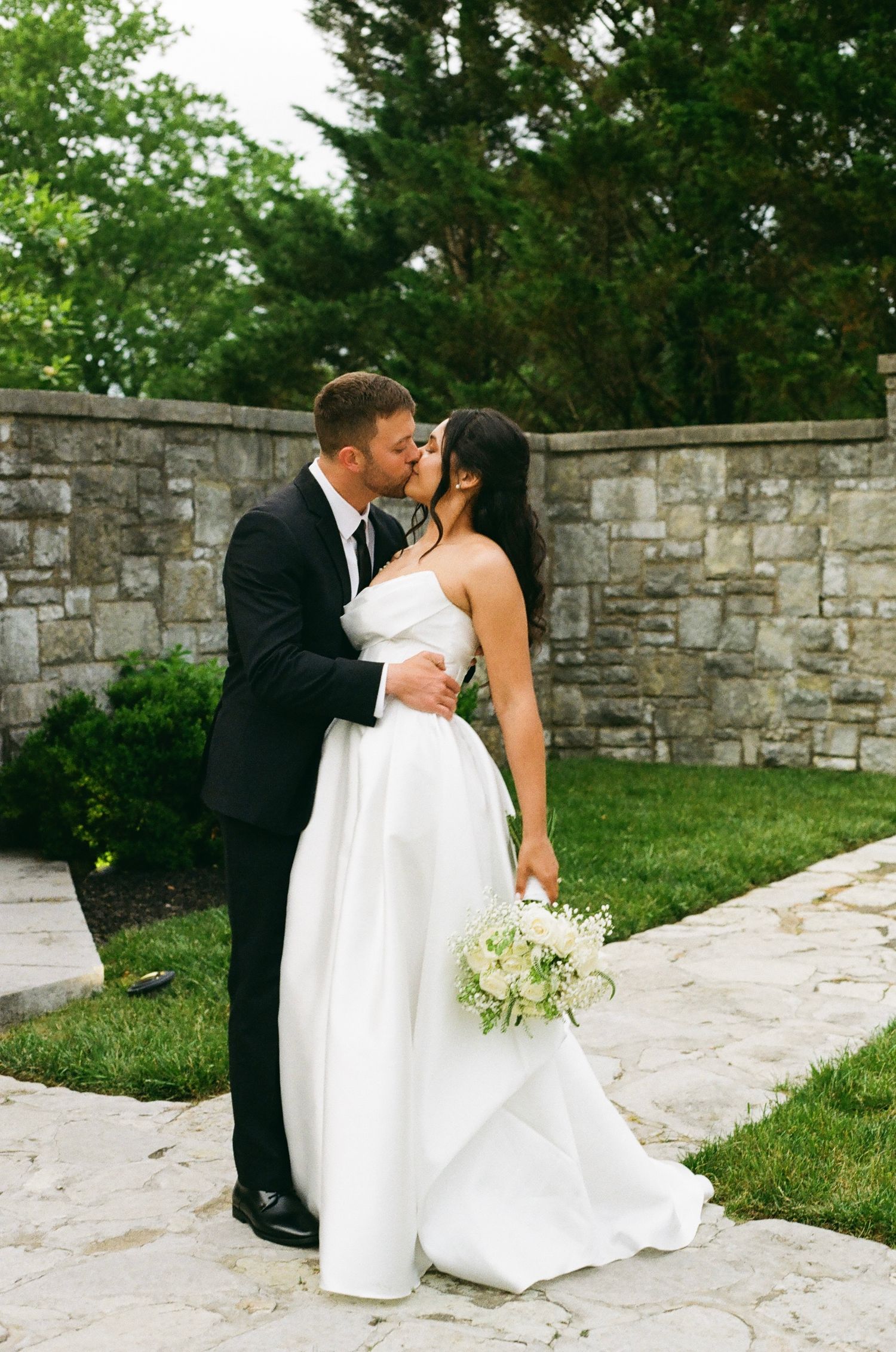 A bride and groom share a romantic kiss outdoors, she holds a white bouquet, stone wall background.