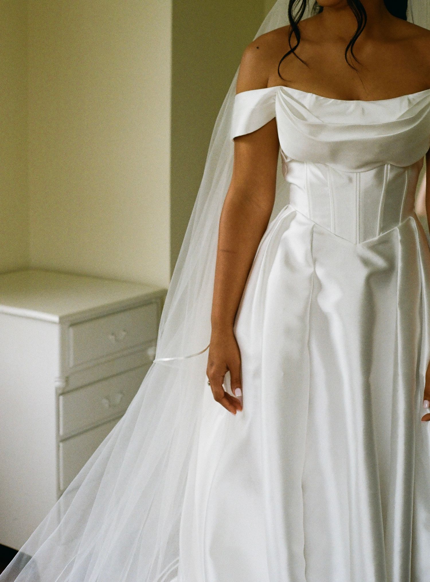 Bride wearing an elegant off-shoulder white satin ball gown with corset bodice and flowing veil in a bridal suite.