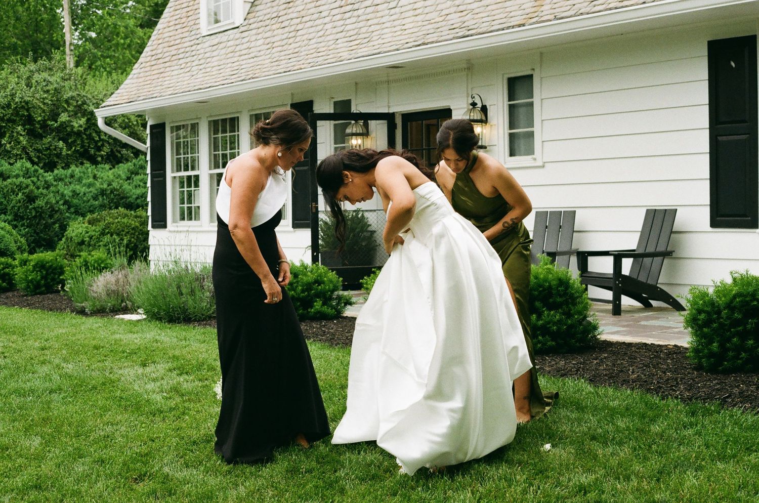 Bride bending over in white gown assisted by two bridesmaids in black dresses on green lawn outside white house.