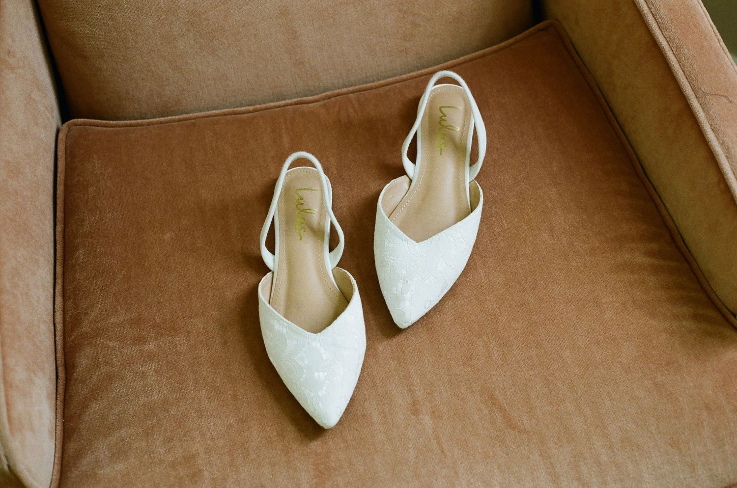 Elegant white pointed-toe slingback flats resting on a velvet peach chair, perfect bridal wedding shoes.