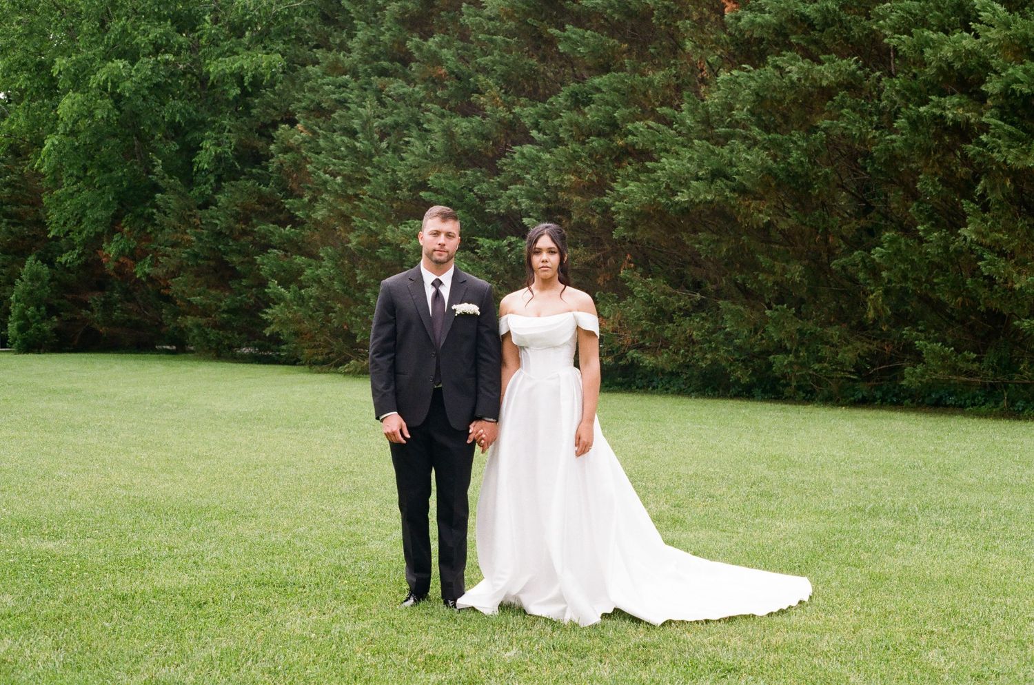 Bride in off-shoulder white gown and groom in black suit stand on green lawn with tall trees behind them.