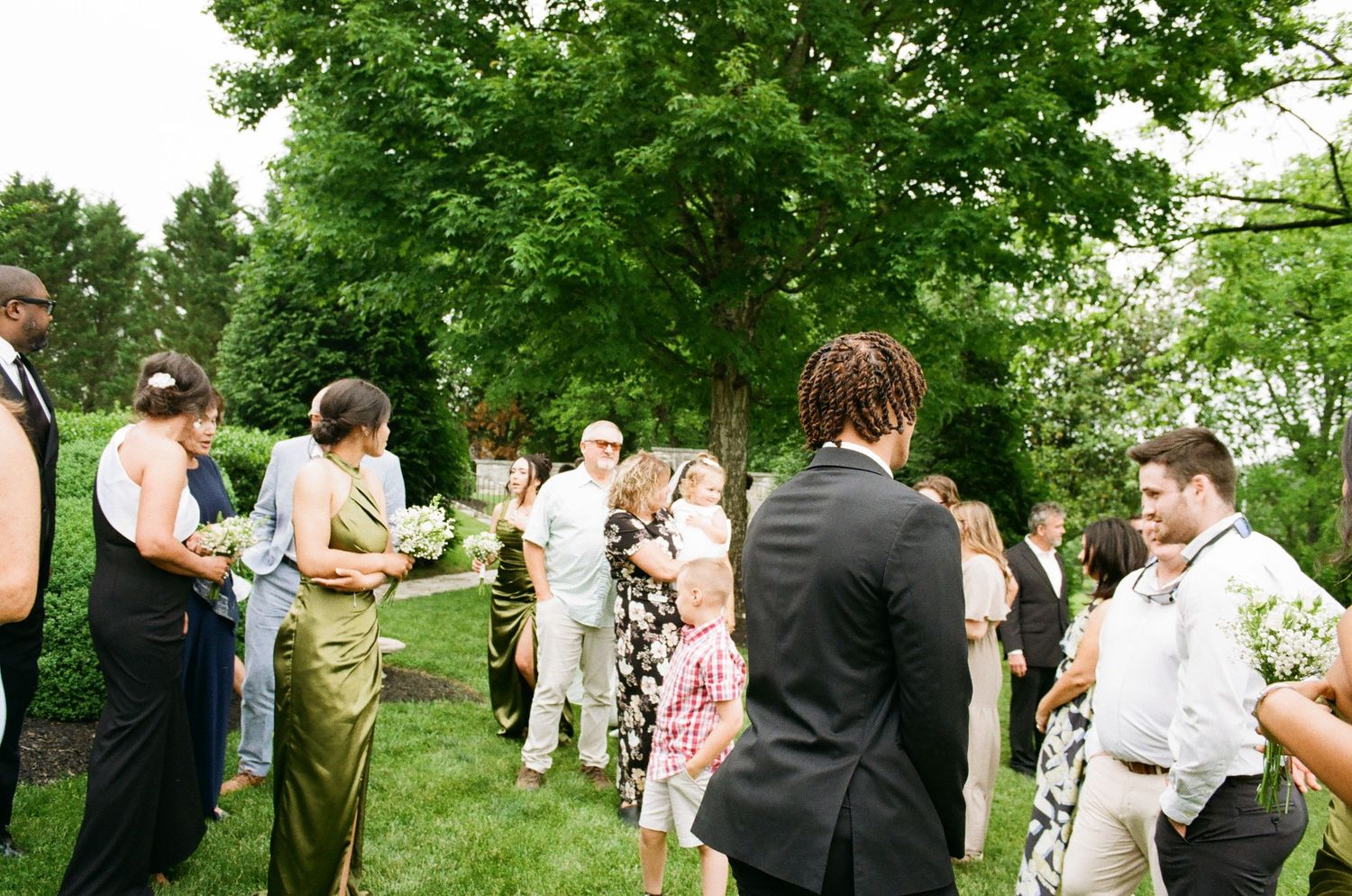 Guests mingle outdoors at an elegant garden wedding ceremony surrounded by lush green trees.