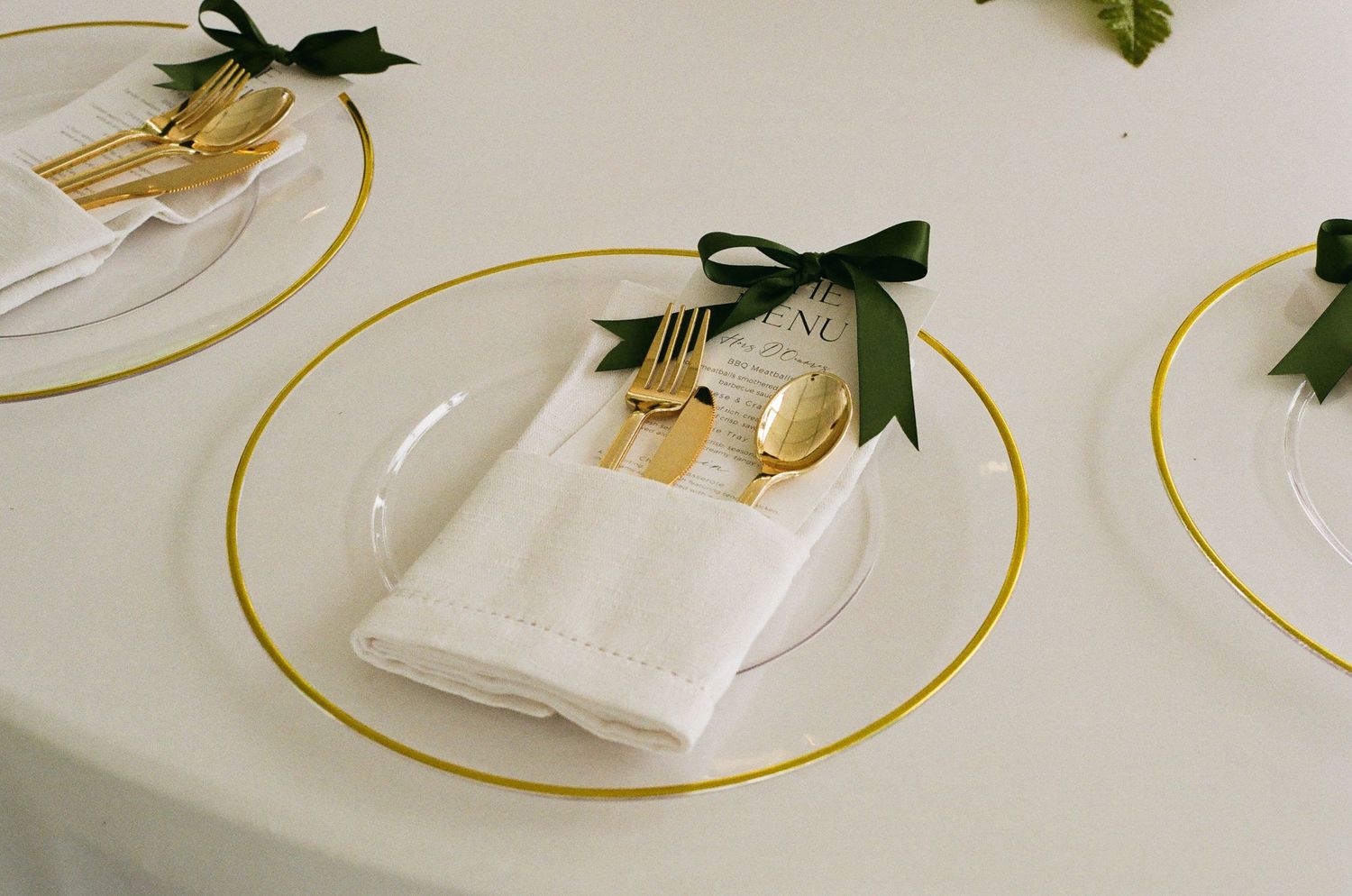 Elegant wedding table setting with gold rimmed plates, gold cutlery, white napkins, and dark green ribbon accents.