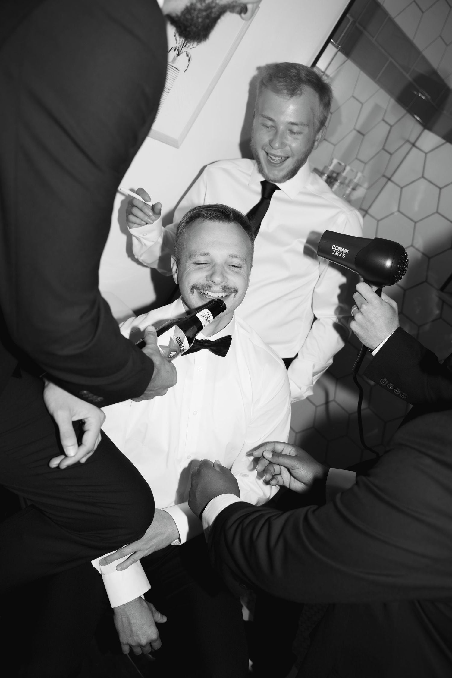 Black and white photo of groomsmen playfully grooming groom with hair dryer and microphone, laughing together.