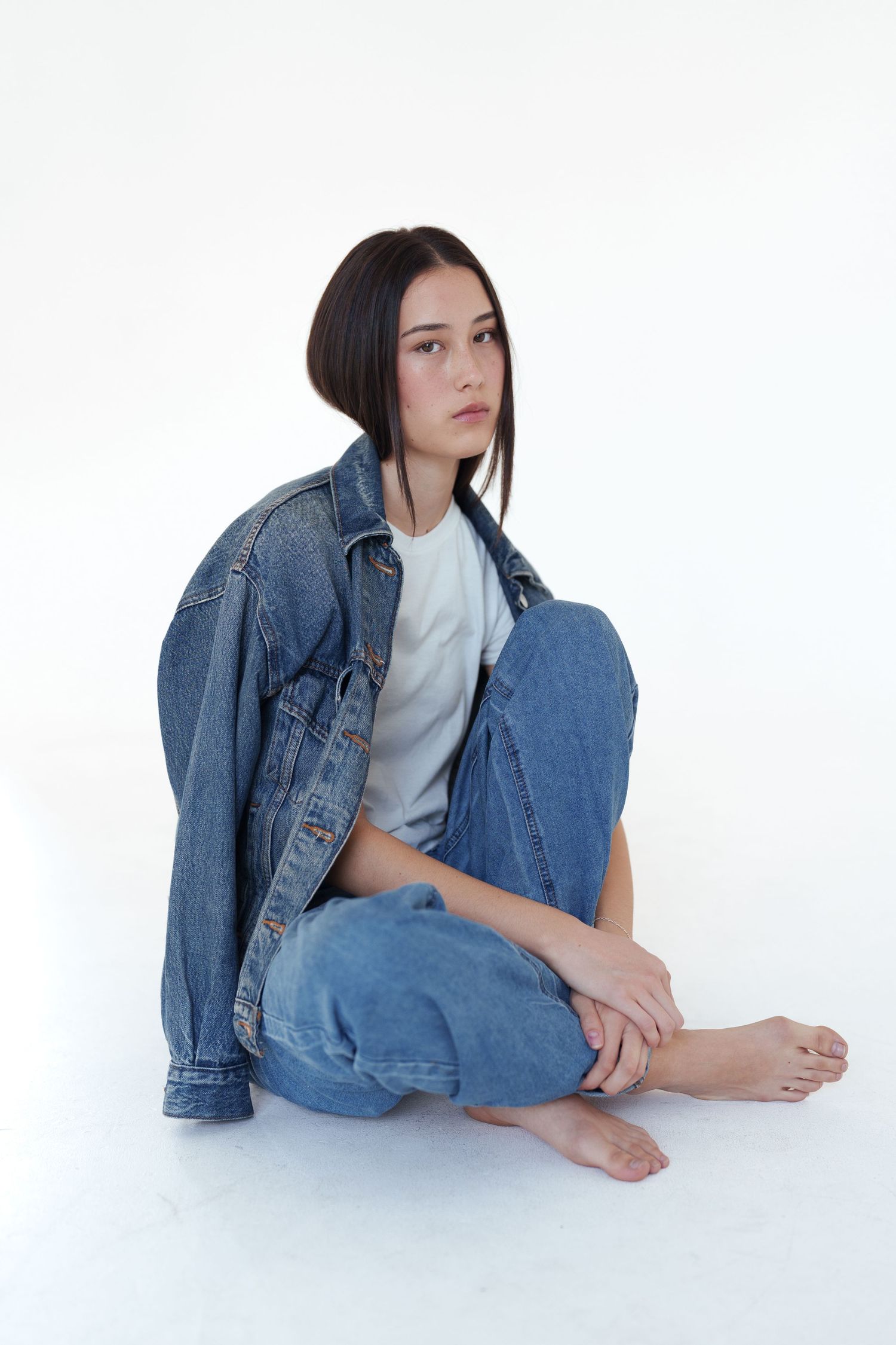 Young woman in oversized denim jacket and jeans sitting barefoot on white background, looking away pensively.