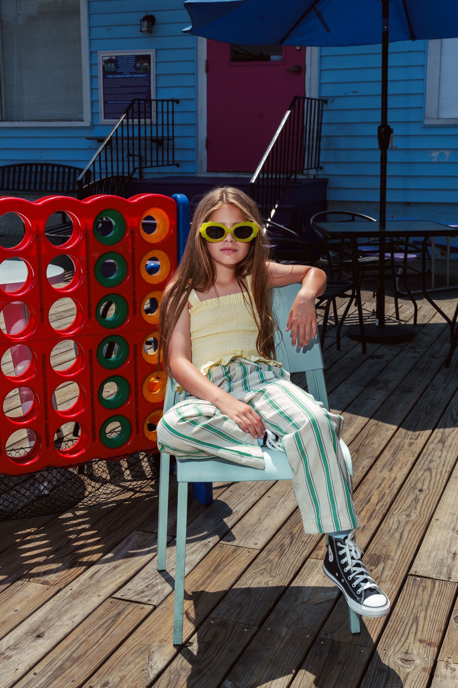 Young child in neon sunglasses lounges on turquoise chair with striped blanket on wooden deck.