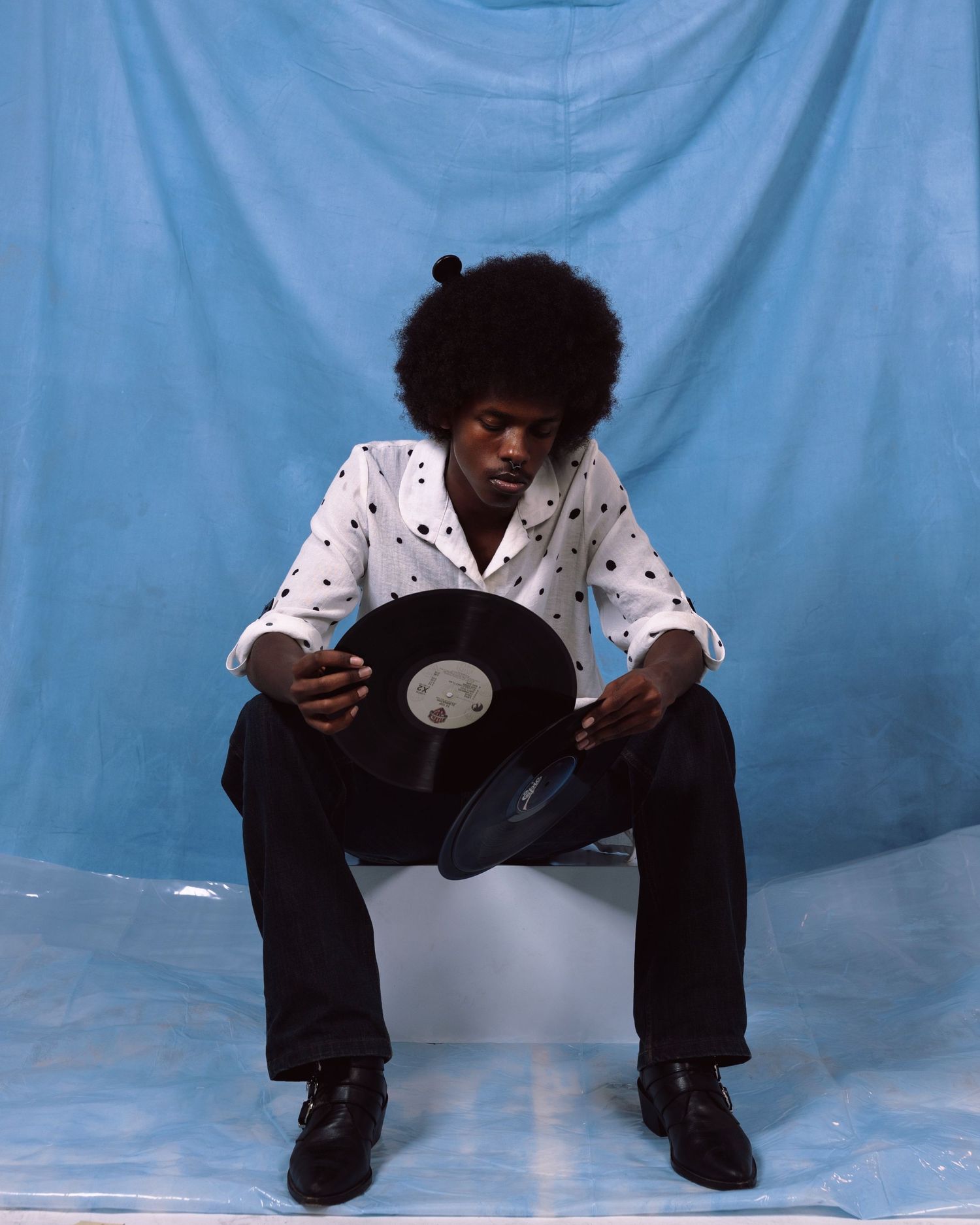 Person in polka dot shirt sits holding vinyl record against blue backdrop with studio lighting.