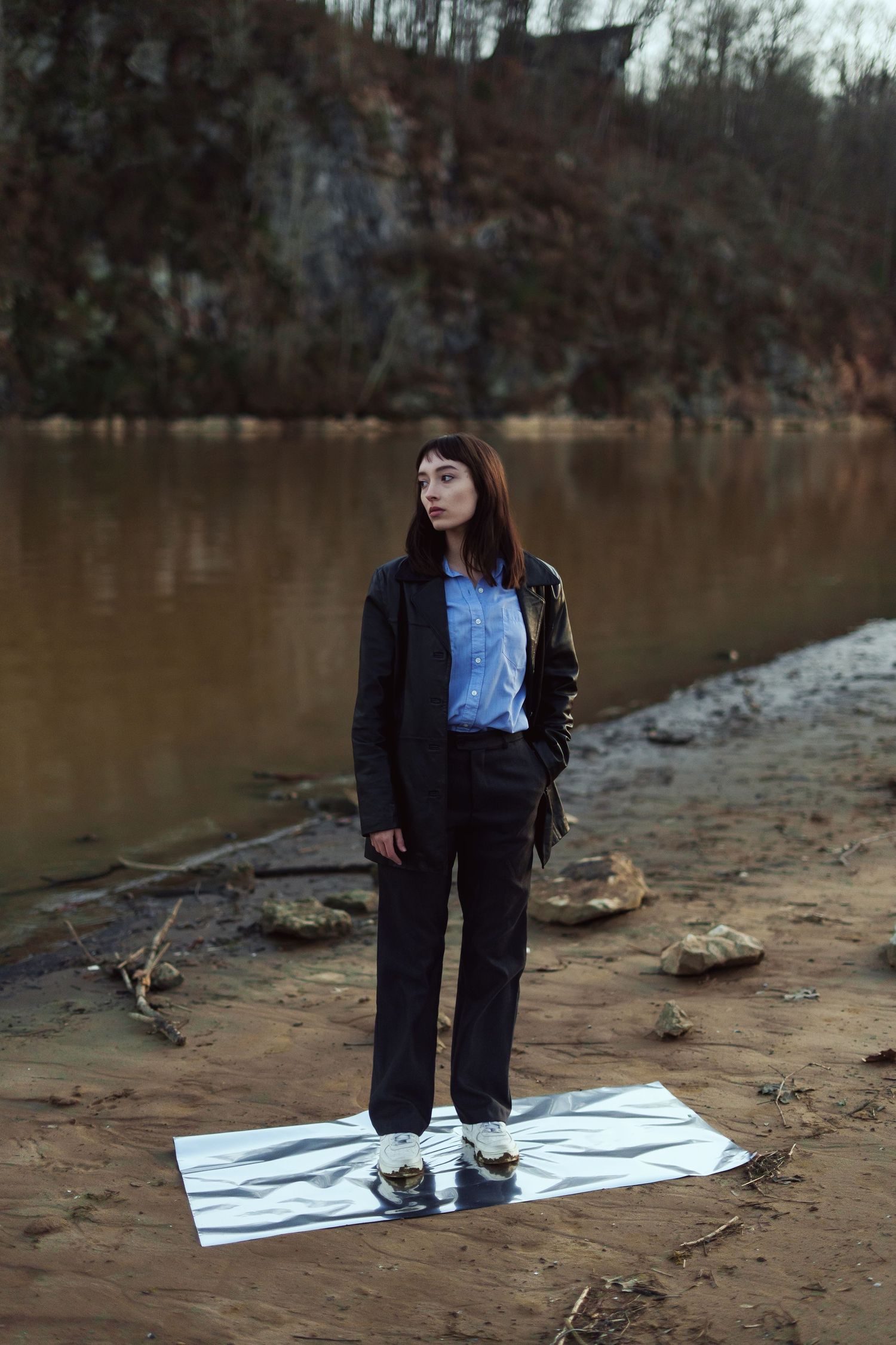A person in a black jacket stands on a white sheet by a brown muddy shoreline near rocky cliffs.
