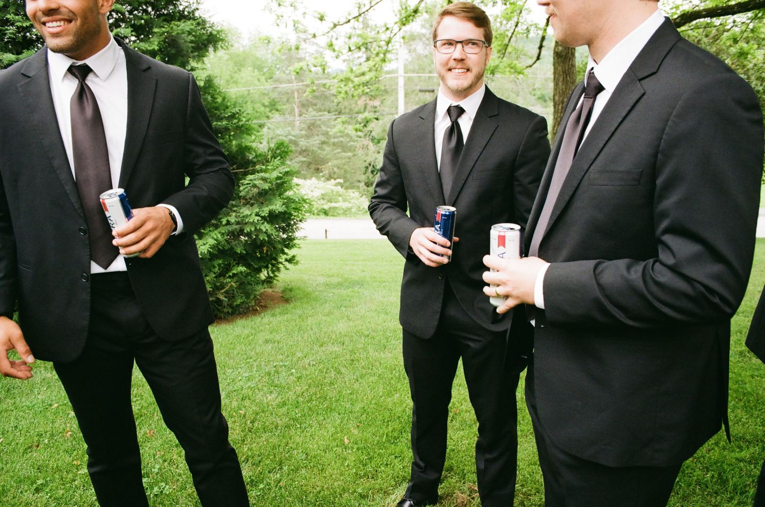 Three groomsmen in black suits and ties laughing outdoors holding drinks at a wedding reception.